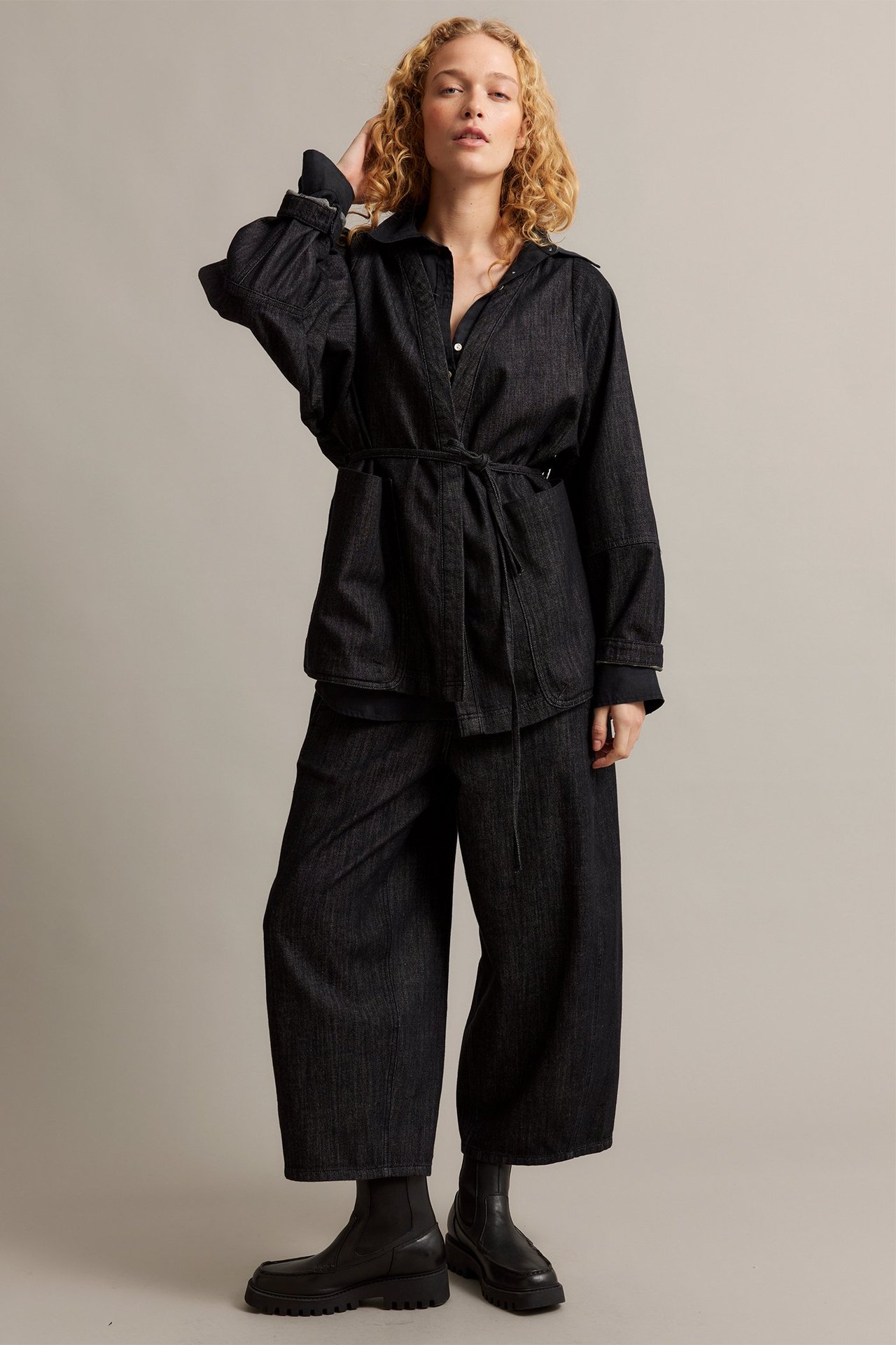 Edestä: Nainen mustassa denimkimonotakissa solmimisvyöllä, leveälahkeisissa housuissa ja mustissa saappaissa.