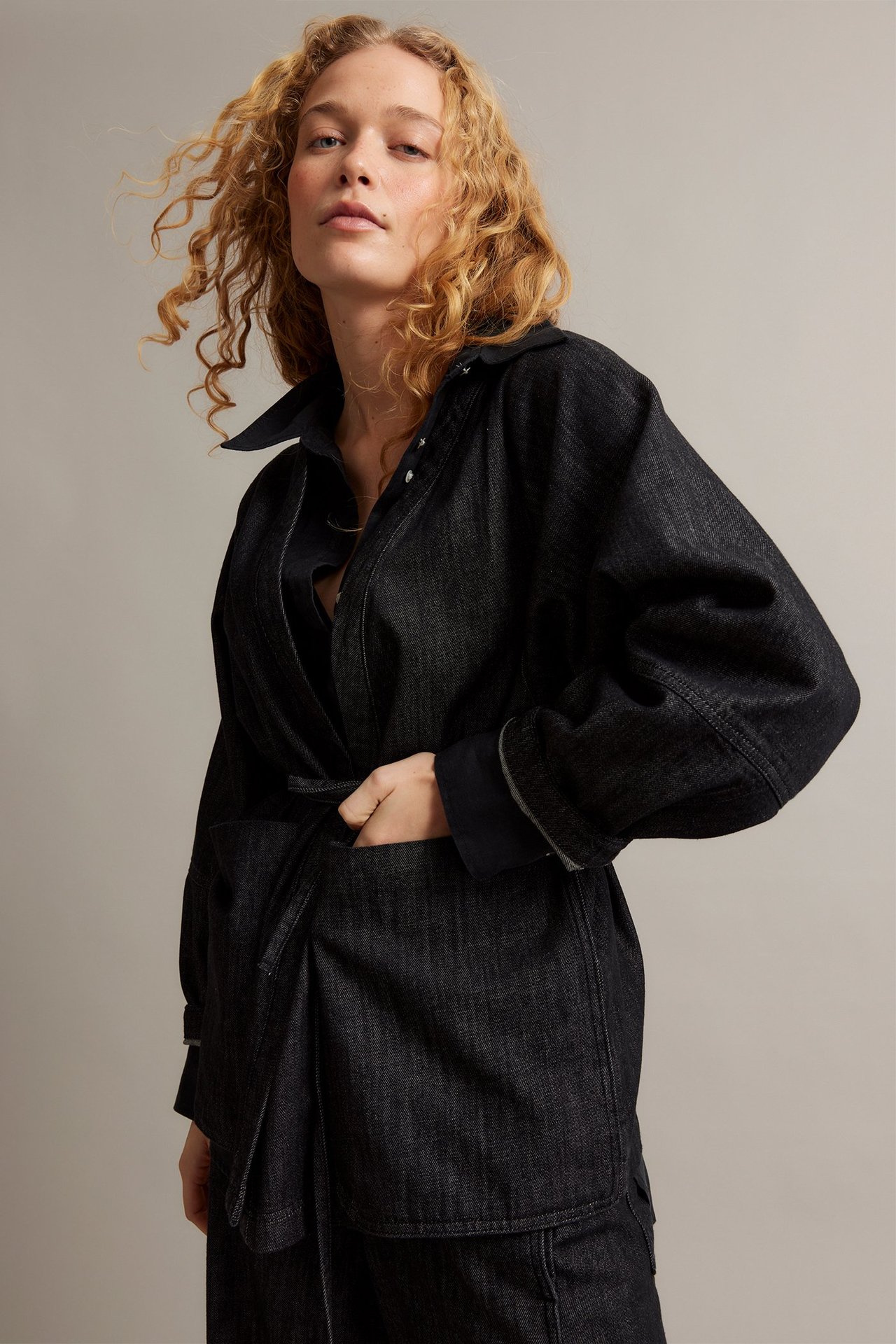 Widok z przodu: Kobieta w czarnej kurtce denim kimono z paskiem i kieszeniami, ręka w kieszeni.