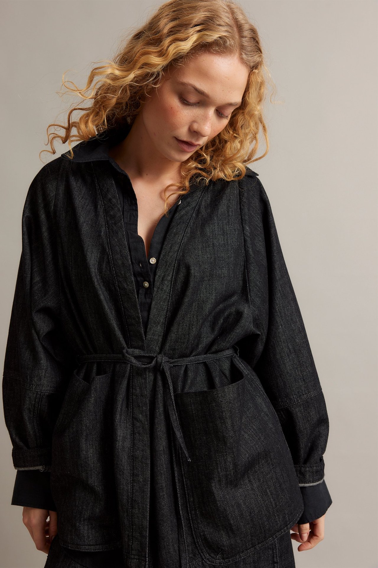 Kobieta w czarnej dżinsowej kurtce kimono z wiązanym paskiem, dużymi kieszeniami, na ciemnej koszuli.