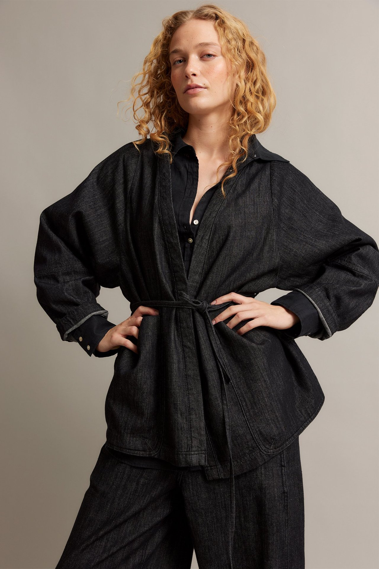 Widok z przodu: Kobieta w czarnej kurtce kimono z denimu z paskiem, na czarnej koszuli i czarnych spodniach.