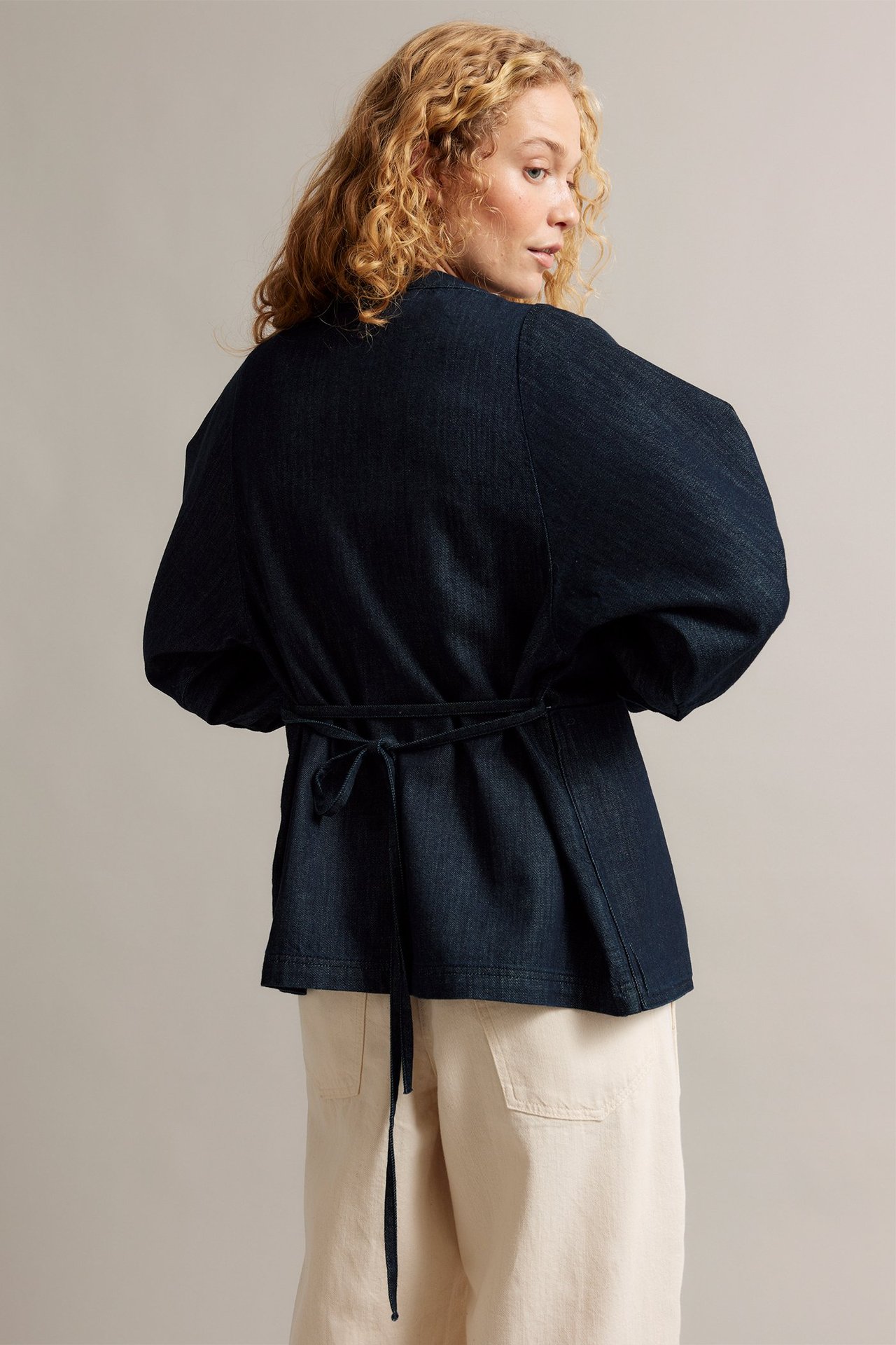 Widok z tyłu kobiety w granatowej dżinsowej kurtce kimono z wiązanym pasem, na beżowych spodniach.