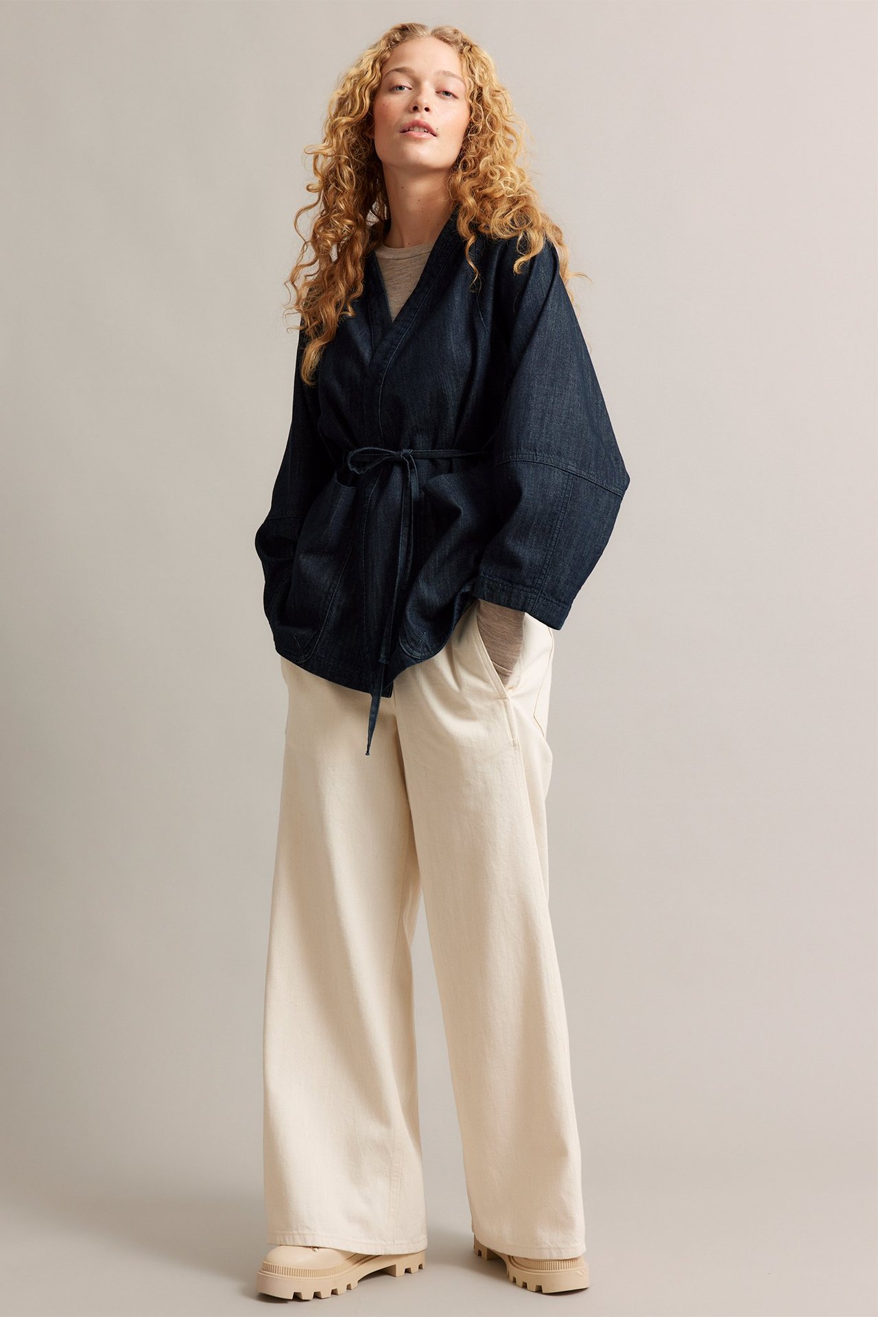 Edestäpäin: Nainen yllään tumma farkku-kimonotakki, vyöllä sidottu, vaalean paidan ja kermanväristen housujen kanssa.