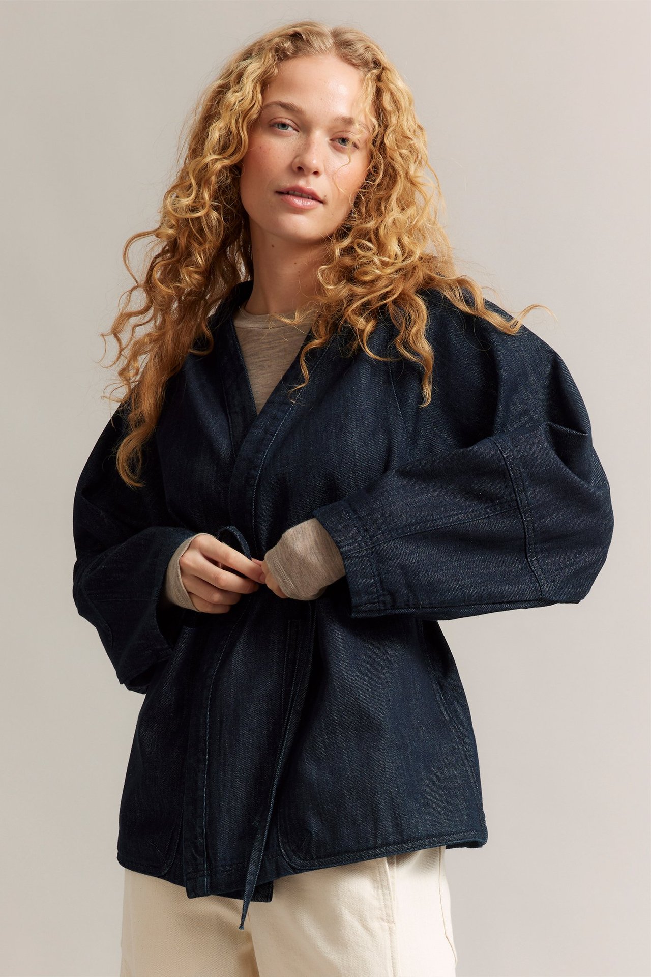 Kobieta w ciemnej dżinsowej kurtce kimono z wiązanym paskiem, jasnym beżowym topie i kremowych spodniach, widok z przodu.