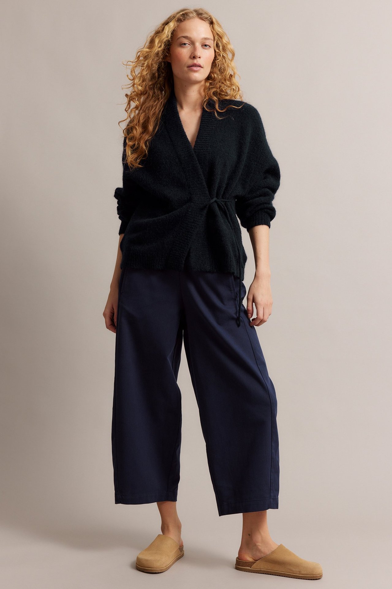 Kvinne i mørk blå ullmiks-omslagscardigan, vide blå bukser og beige slip-on-tøfler.