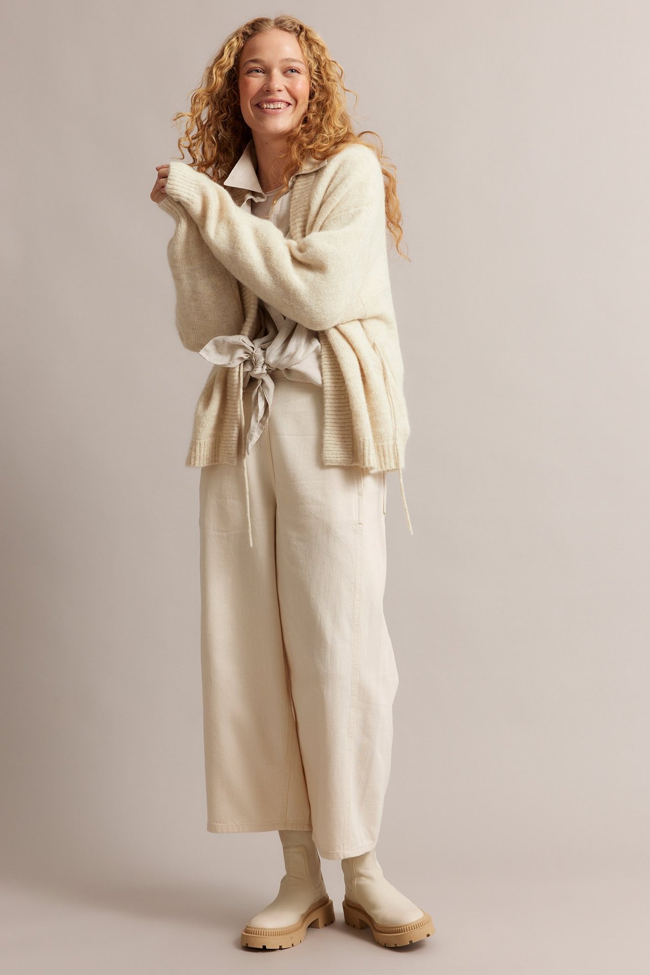 Forfra: kvinne i lys beige ullmiks-cardigan med knytebånd, lys skjorte, vide bukser og støvler.