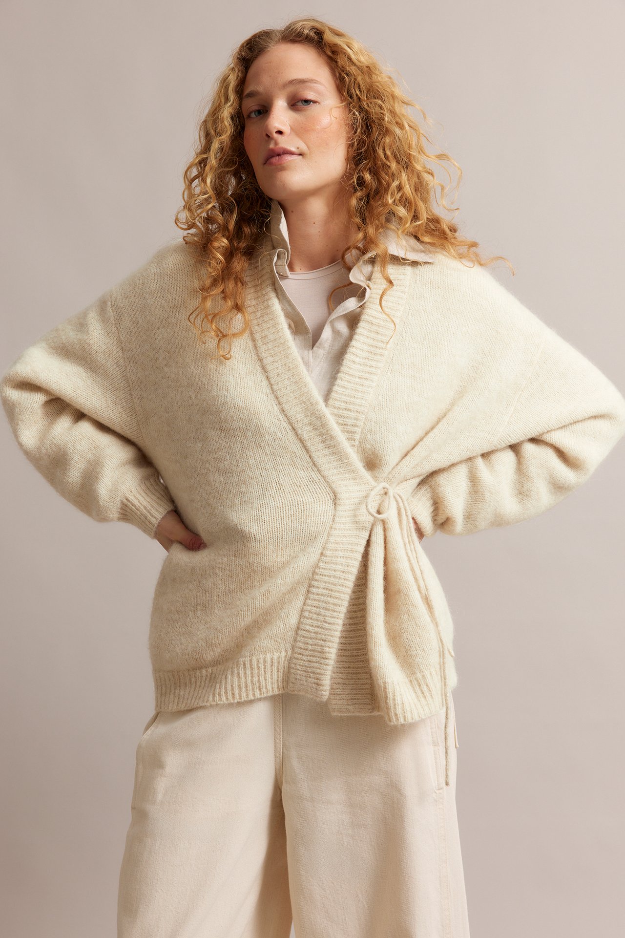 Naisen etukuva: Vaaleabeige villasekoitekardigaani solmimisnauhoilla, paidan ja housujen päällä.