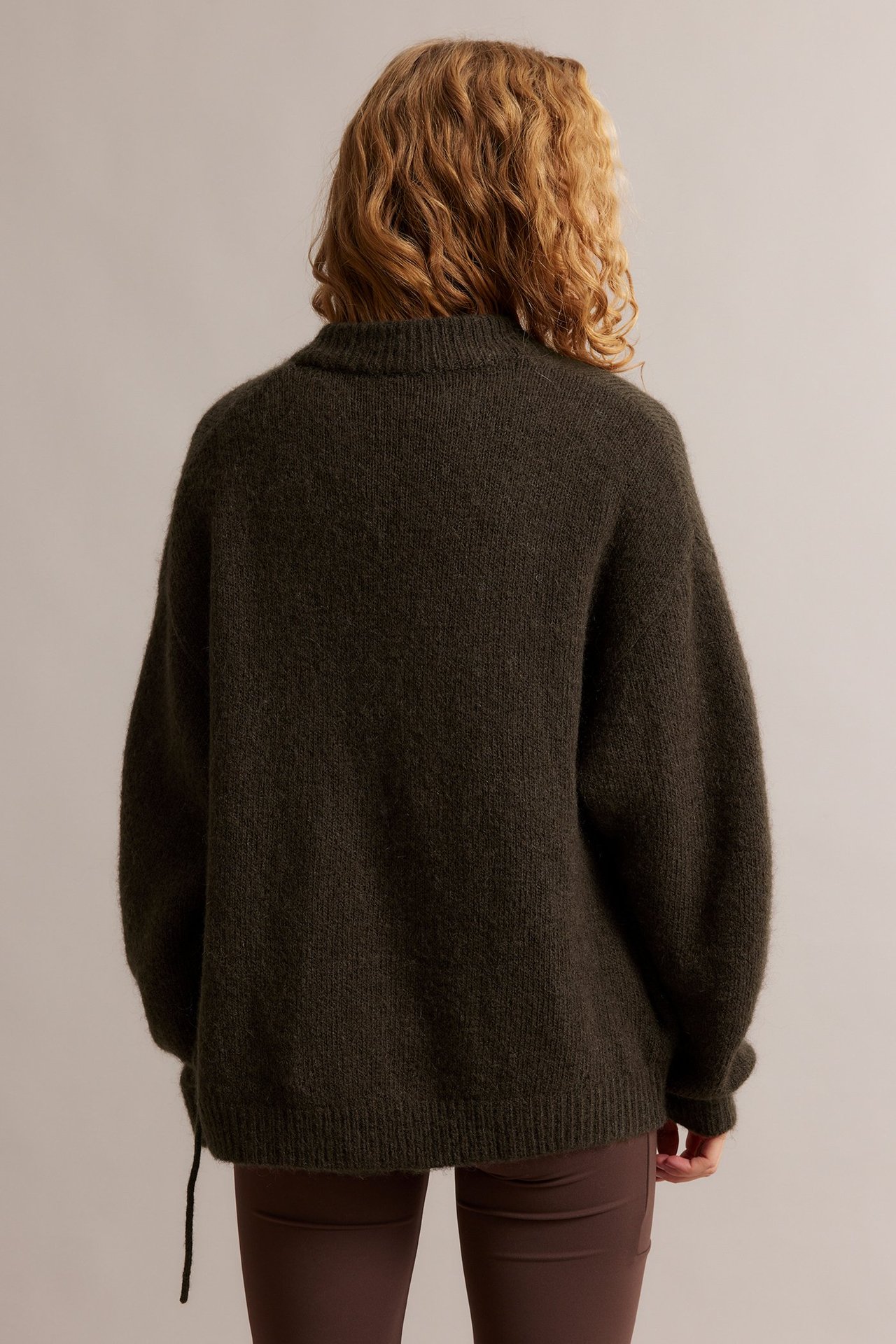 Naisen takakuva tummanruskeassa villasekoitekardigaanissa, jossa on solmimisnauha ruskeiden housujen päällä.