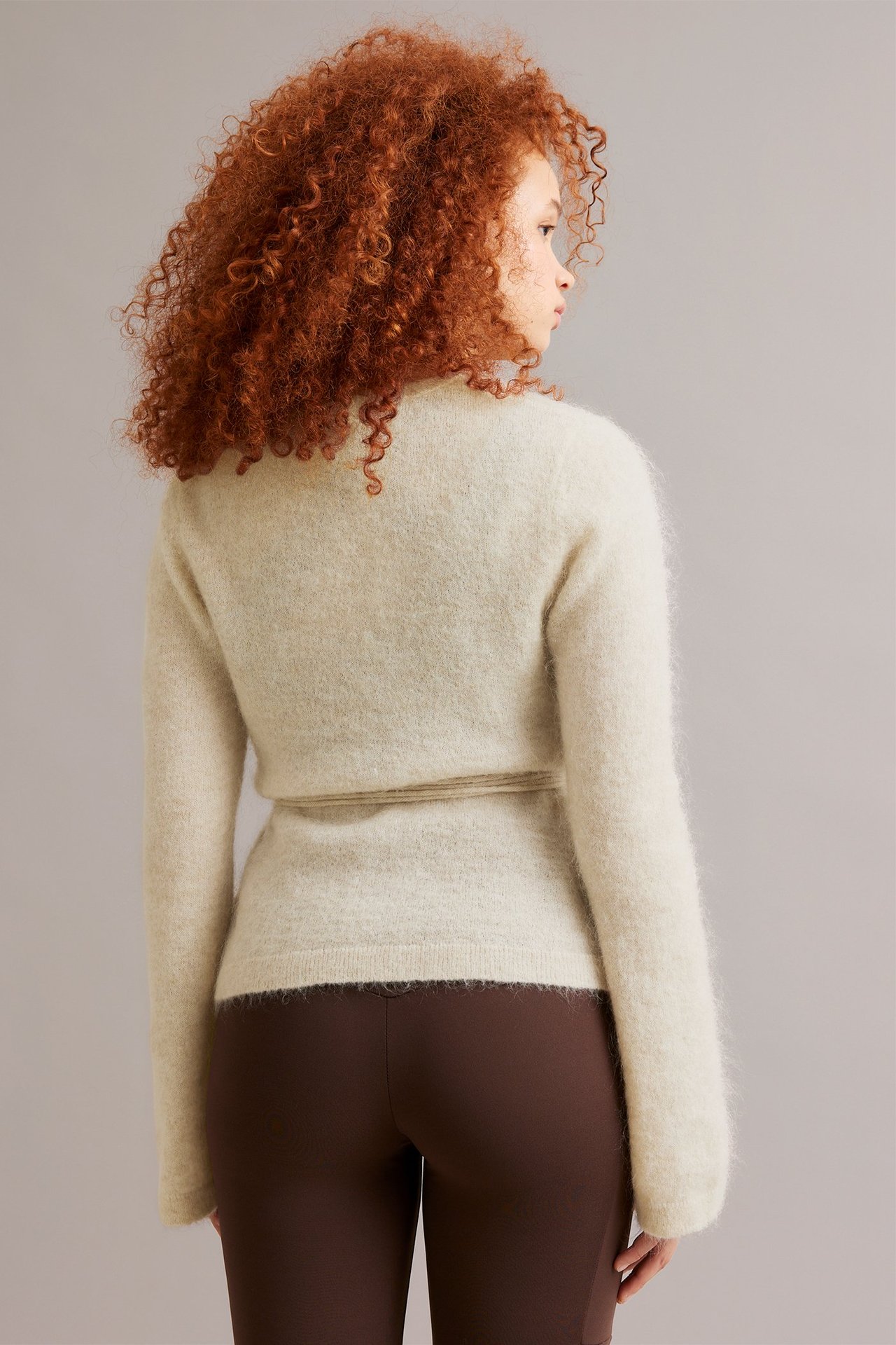 Nainen takaa, yllään vaalea beige villasekoite kietaisuneuletakki vyöllä ja ruskeat leggingsit.