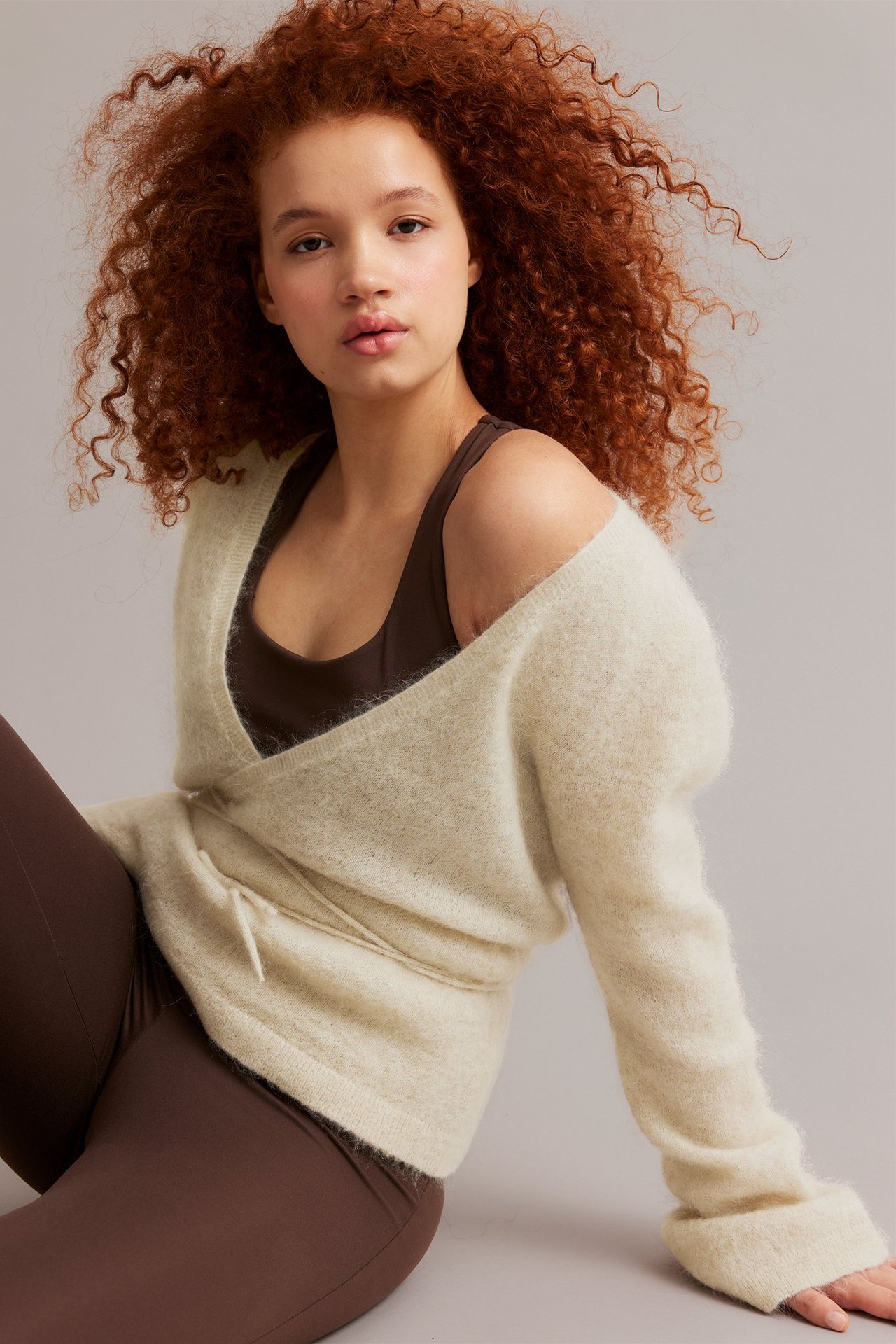 Nainen yllään vaaleabeige kietaisuneuletakki, vyötäröltä sidottu, ruskean rintaliivin ja leggingsien päällä.