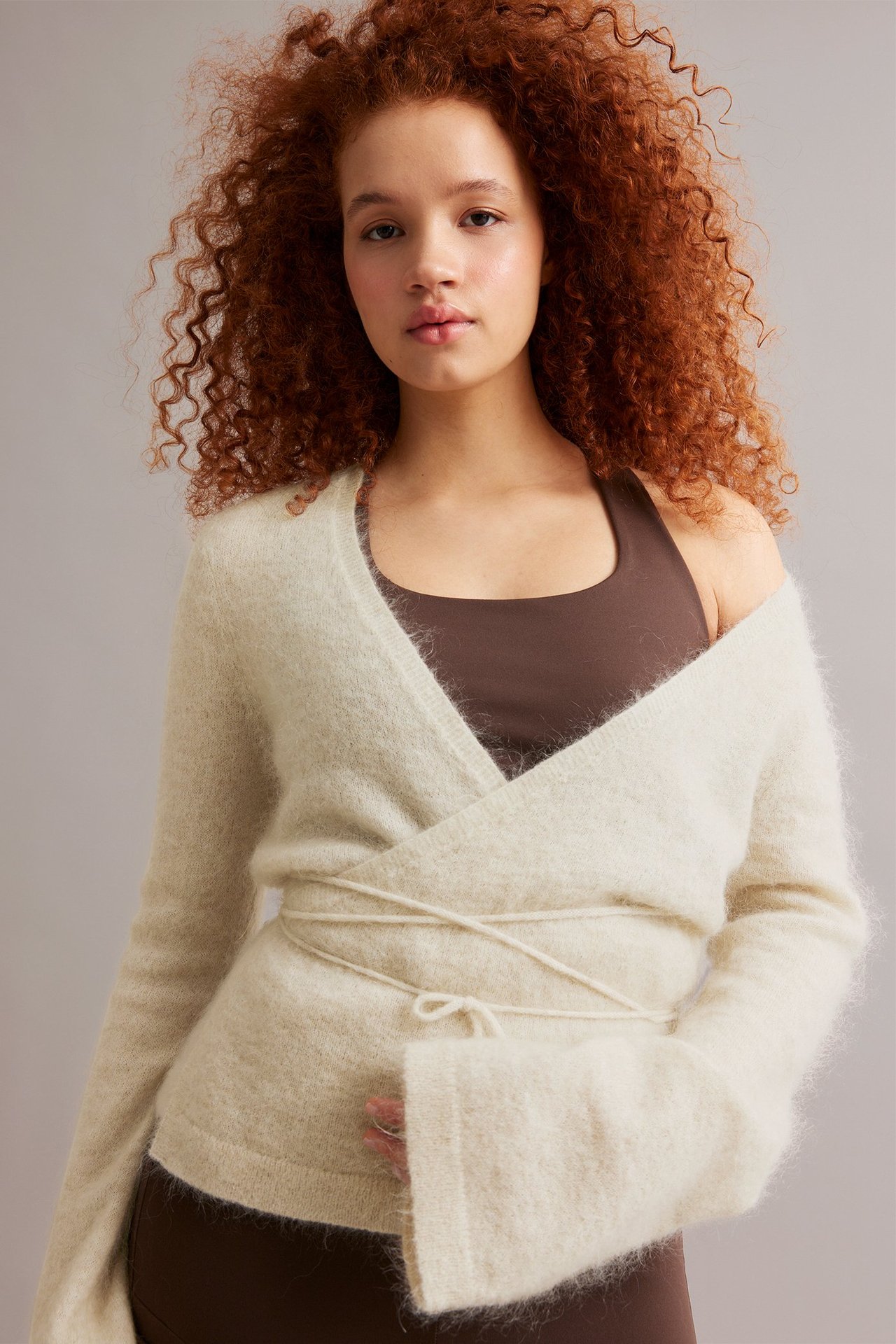 Nainen edestäpäin yllään vaaleabeige, solmittava, ohut villasekoitekietaisuneule ruskean topin päällä.