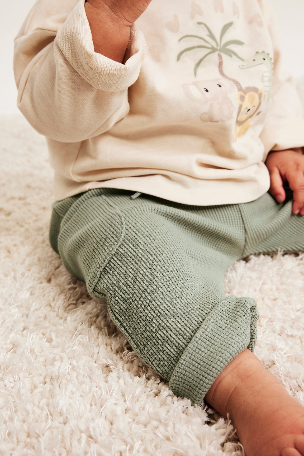 Närbild på ett baby i mörkgröna våfflade byxor med dragsko och en beige topp med djurmönster.
