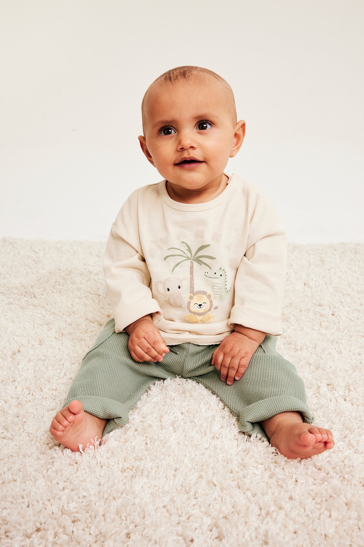 Frontvy av en baby som sitter, klädd i mörkgröna våfflade byxor och en beige långärmad tröja med djurmönster.