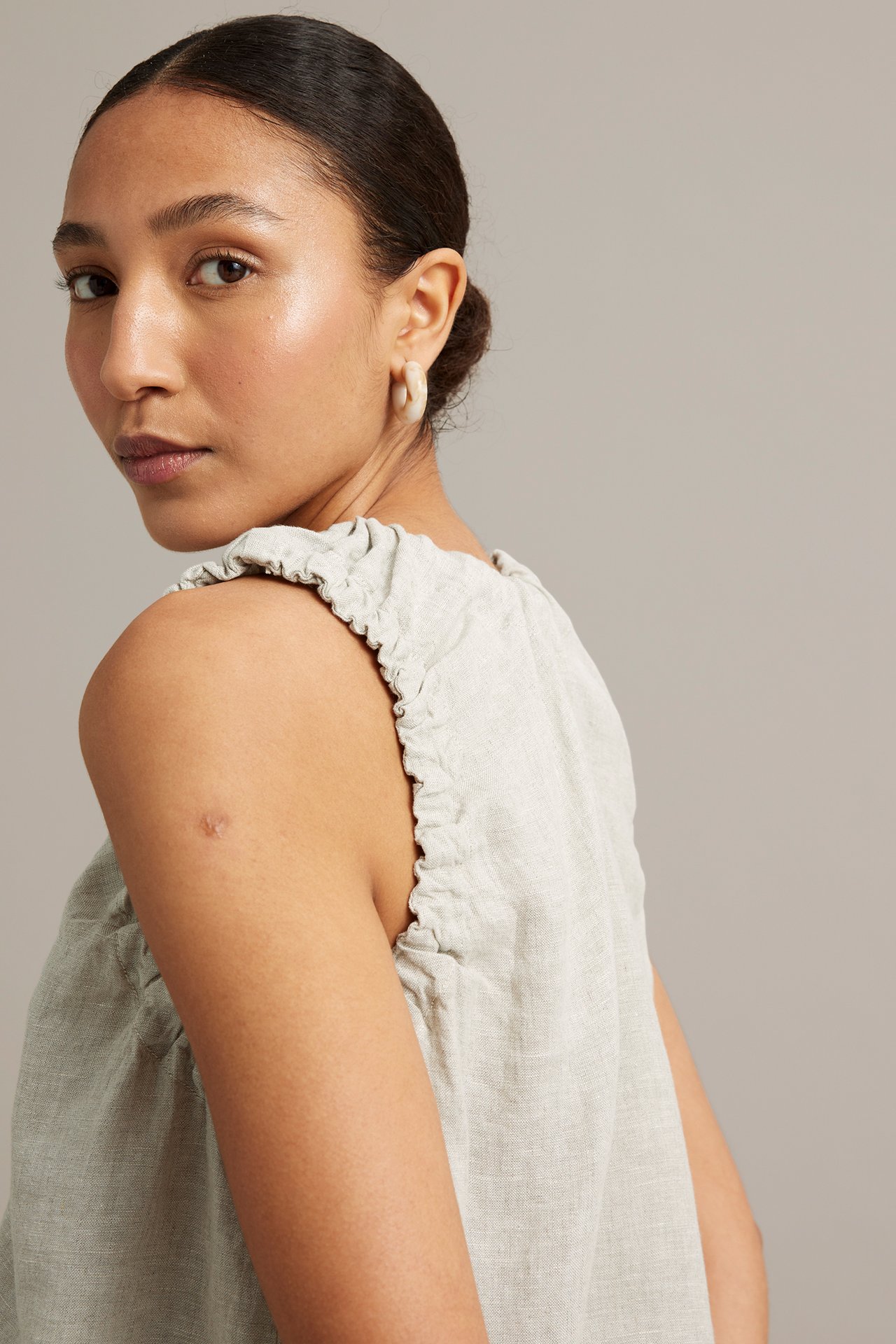 Närbild bakifrån av kvinna i ljusbeige linnetopp med rynkade axeldetaljer och ljusa ringörhängen.