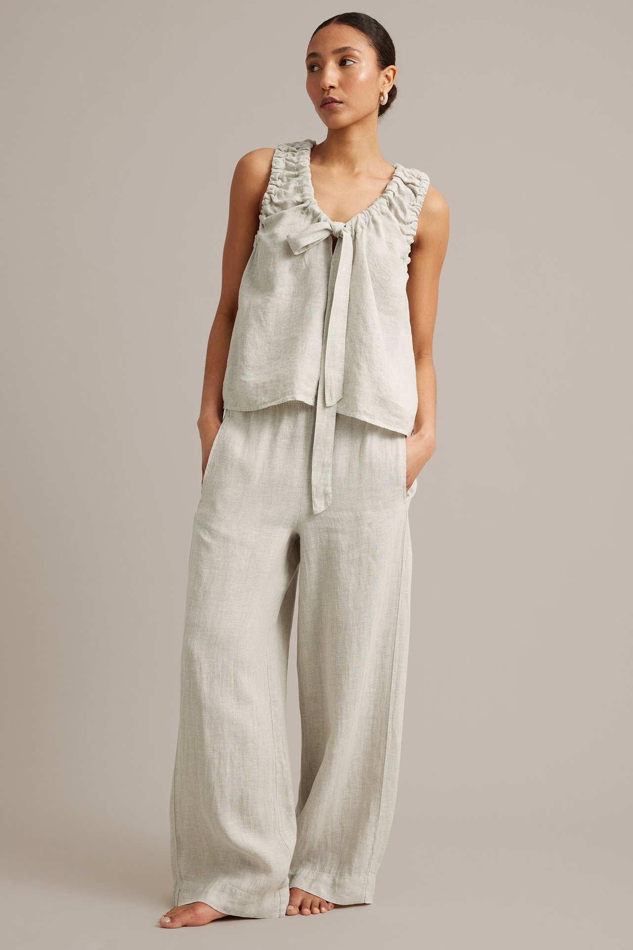 Framifrån, kvinna i lös ljusbeige linnetopp med knytband i halsringningen och matchande vida linnebyxor.