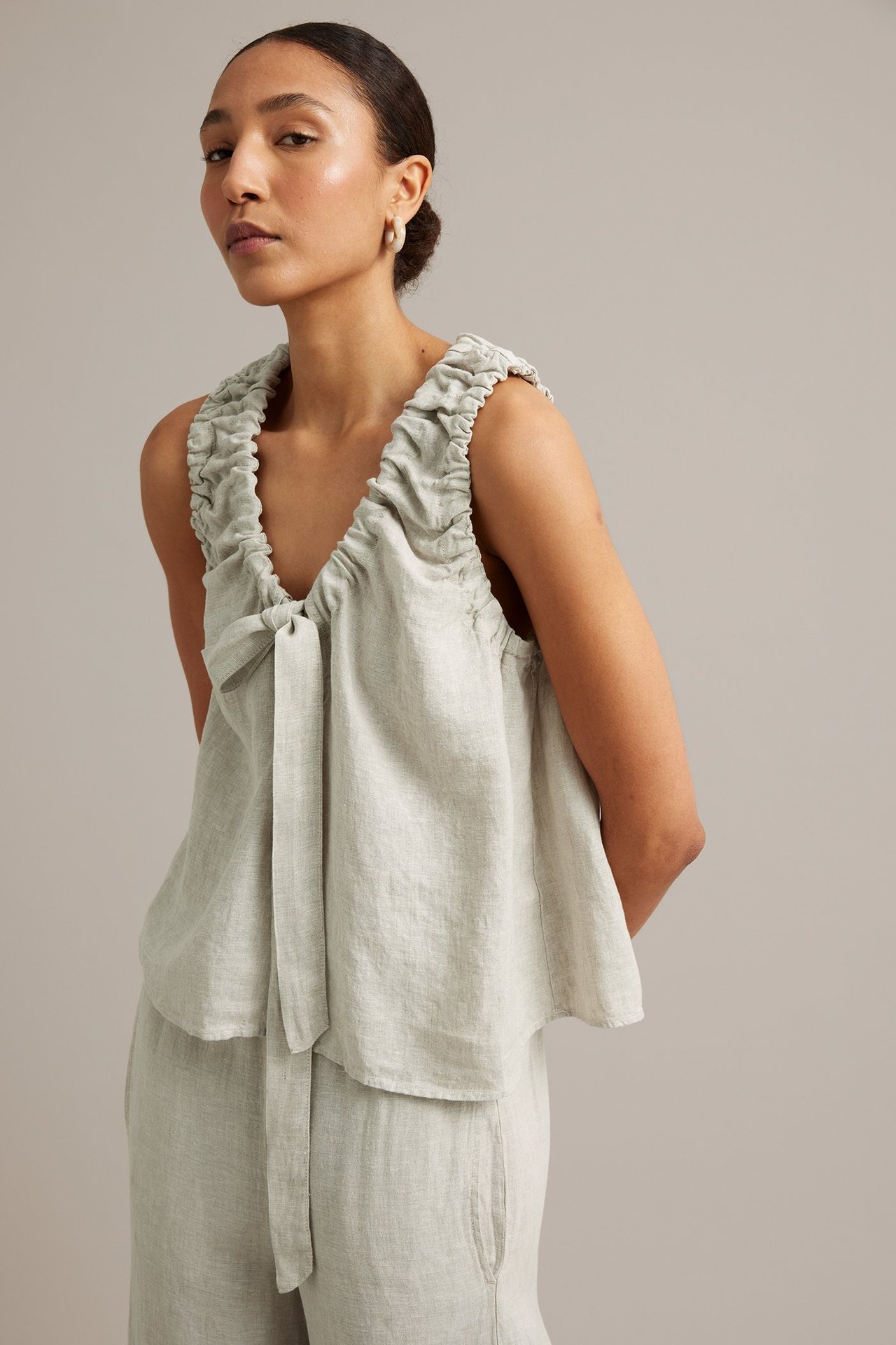 Framsida: Kvinna i lös beige linnetopp med rynkad V-ringning, knytband fram och matchande byxor.