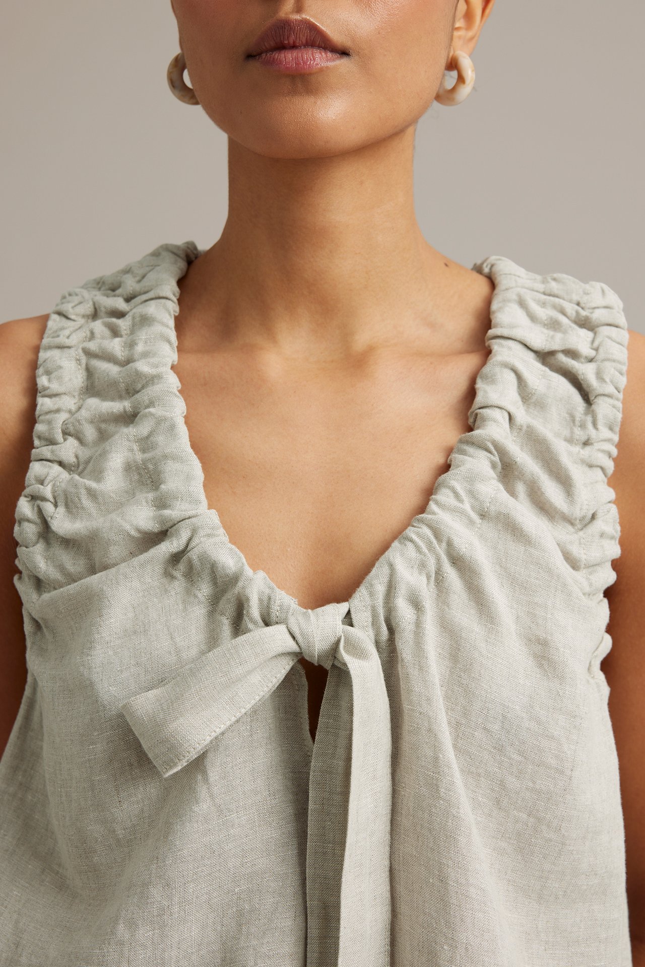 Närbild av en kvinna i beige linnetopp med rynkad ringning och knytband framtill, plus örhängen.