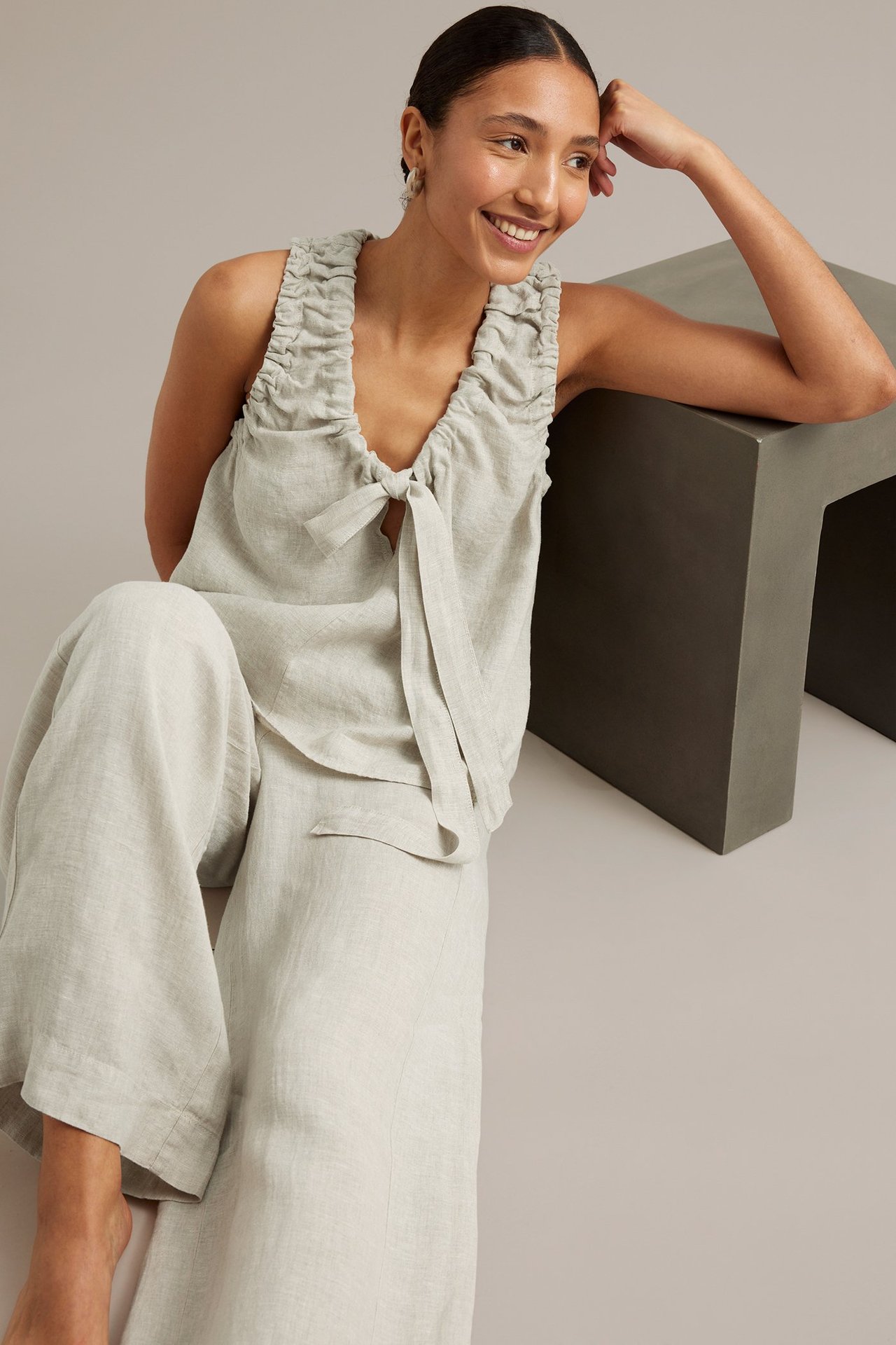 Framifrån, leende kvinna bär beige linnetopp med dragsko och knytband samt matchande vida linnebyxor.