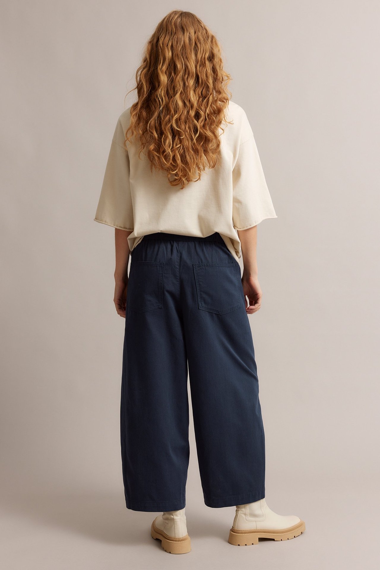 Z tyłu: kobieta w granatowych spodniach pull-on z szerokimi nogawkami, tylnymi kieszeniami, kremowym t-shircie i beżowych botkach.