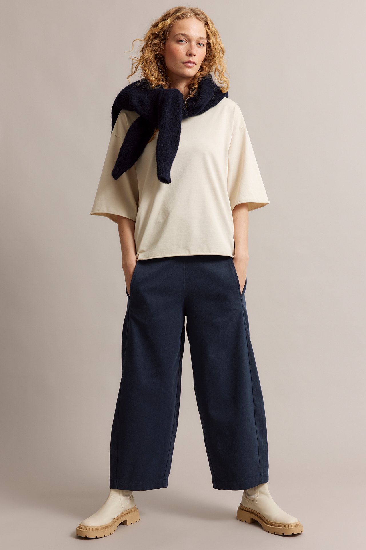 Edestä näkymä naisesta tummansinisissä pull-on-housuissa, beigessä topissa ja tummansininen neule kaulalla.