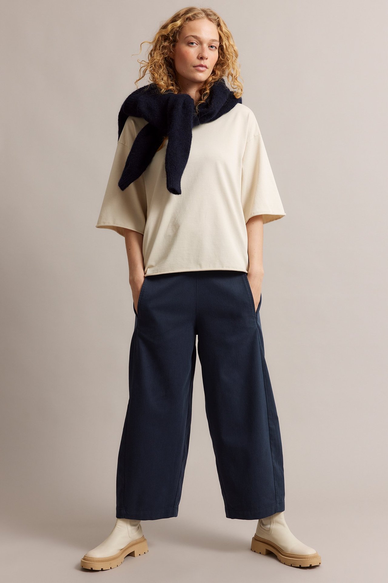 Edestä näkymä naisesta tummansinisissä pull-on-housuissa, beigessä topissa ja tummansininen neule kaulalla.