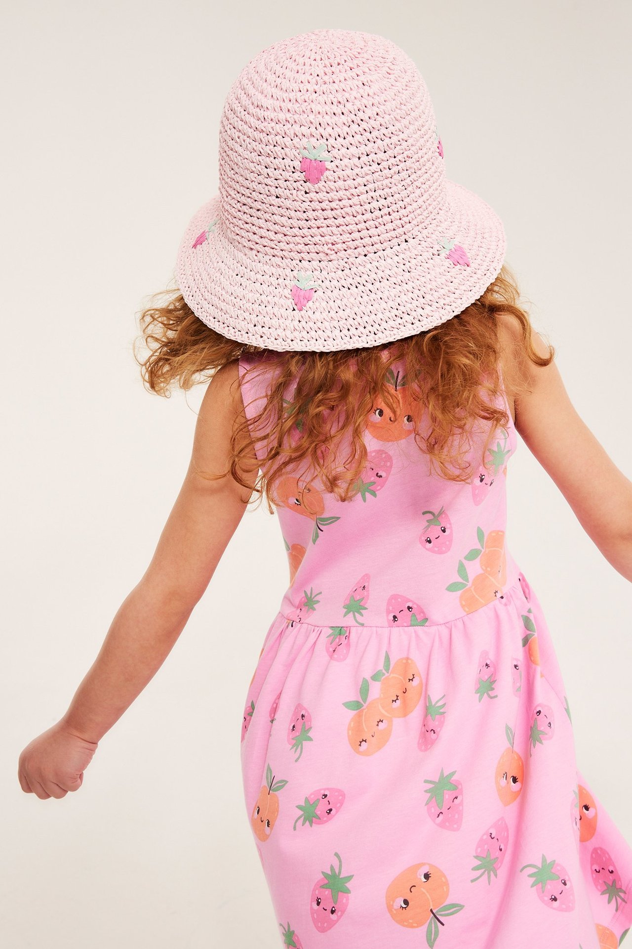 Bakifrån på barn med rosa stråhatt med broderade jordgubbar och en rosa klänning med fruktmönster.