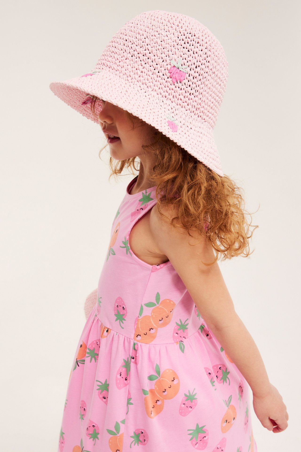 Sidovy av ett barn i rosa stråhatt med jordgubbsapplikationer och en rosa klänning med fruktmönster.