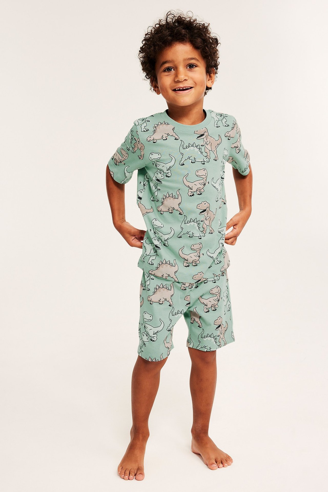Edestä kuvattu lapsi vihreässä lyhythihaisessa dinosauruskuvioisessa puuvillapyjamassa.
