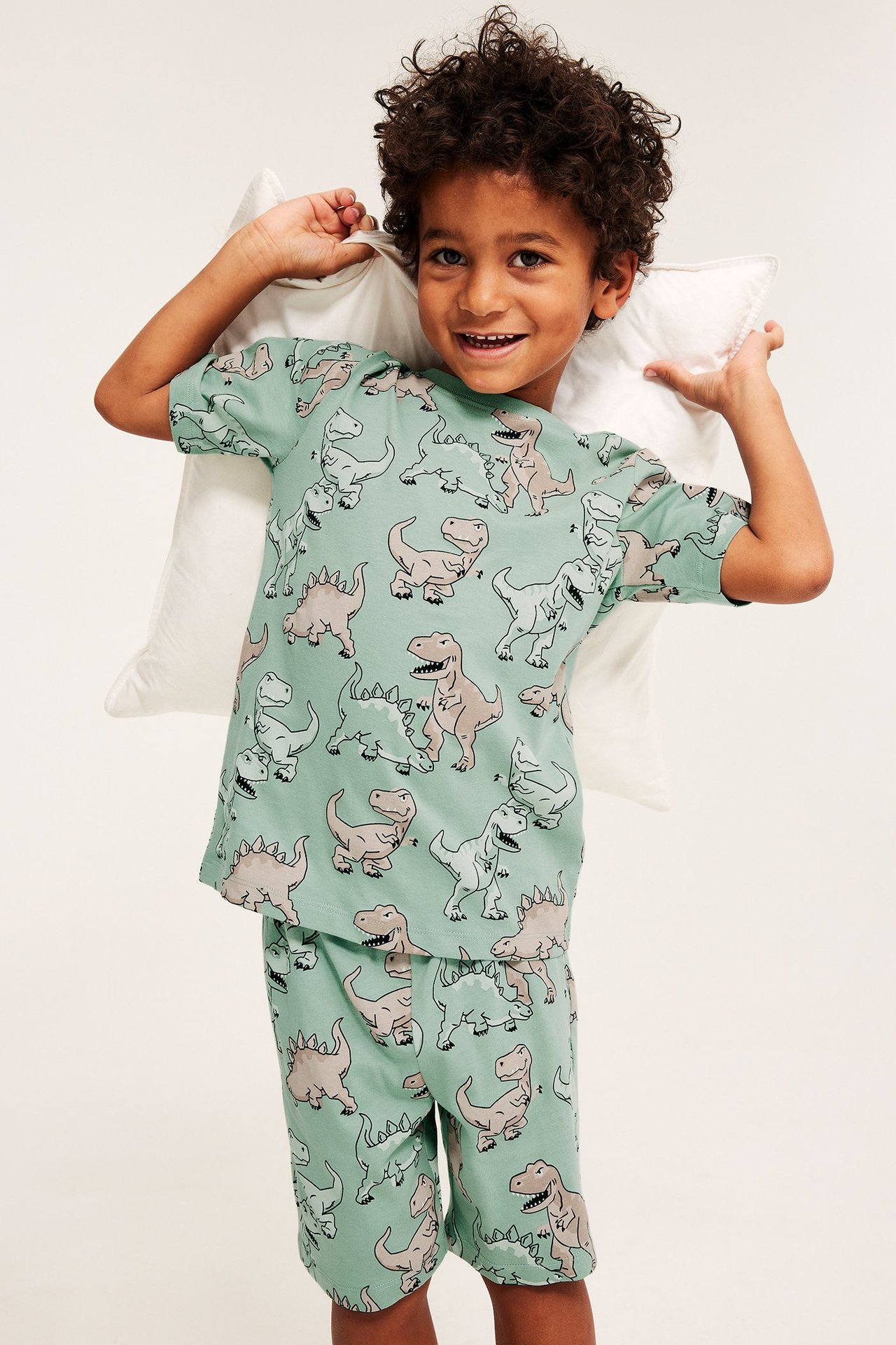 Przód: dziecko w zielonej bawełnianej piżamie w dinozaury, trzyma białą poduszkę.