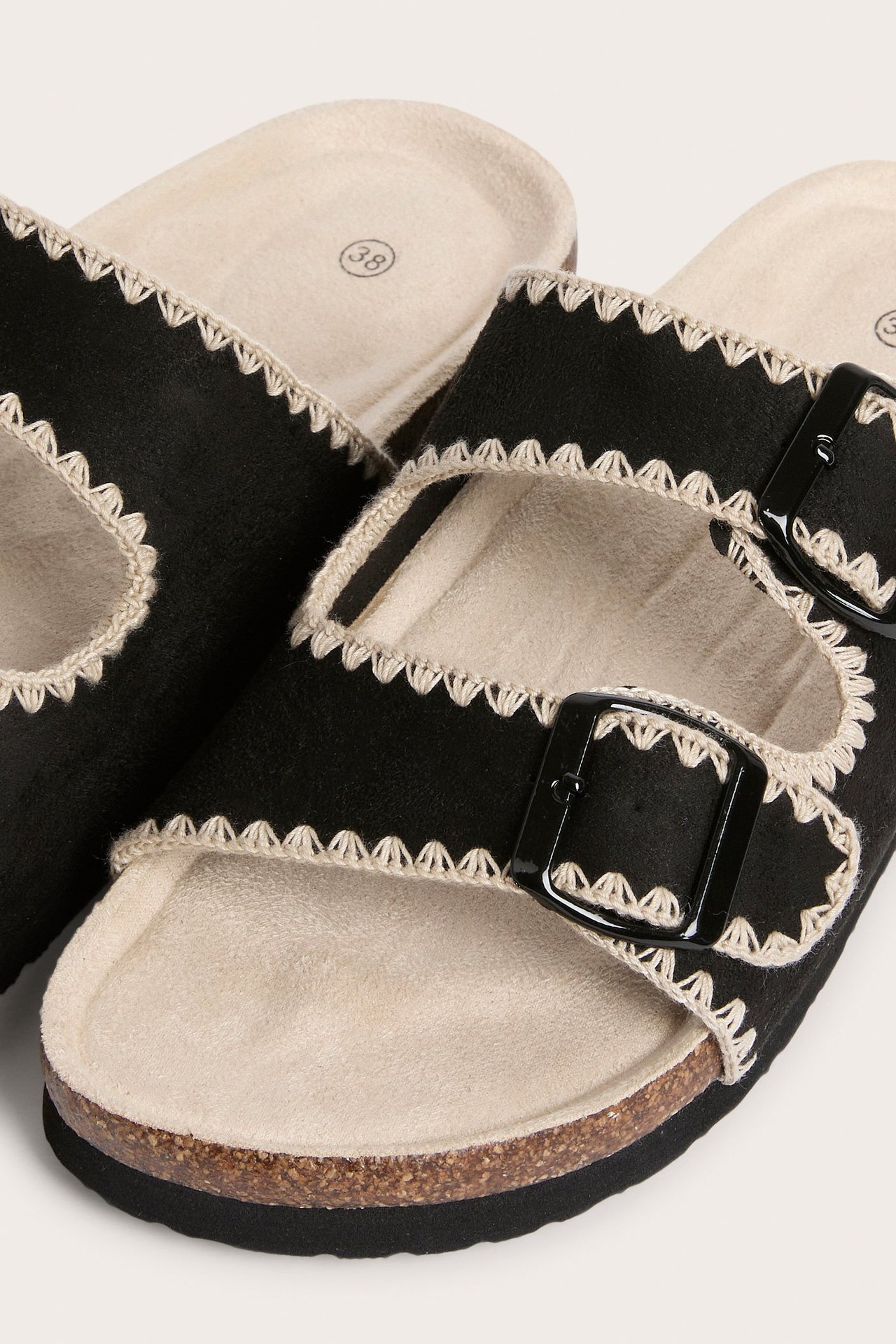 Zbliżenie dwóch czarnych sandałów z podwójnymi paskami, czarnymi klamrami, jasnym falowanym szwem i beżową wkładką.