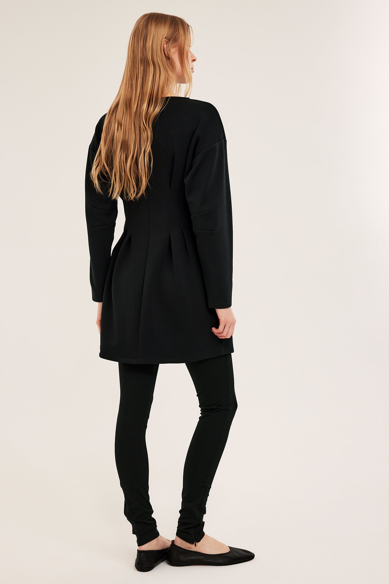 Ryggvy av kvinna i svart jerseyklänning med intagen midja, svarta leggings och svarta ballerinaskor.