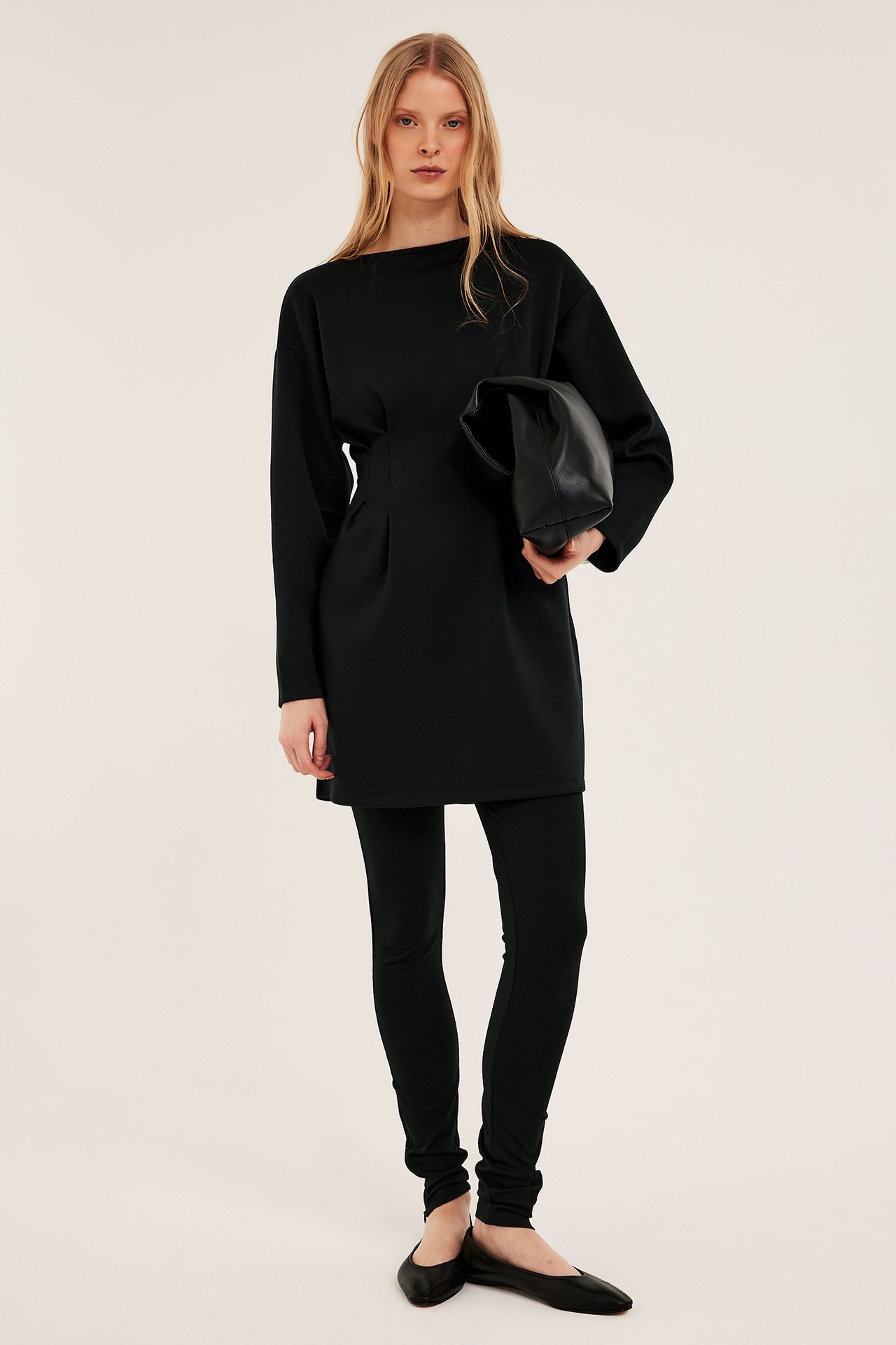 Frontalvisning: Kvinne i svart jerseykjole med markert midje, svarte leggings, flate sko, holder svart clutchveske.