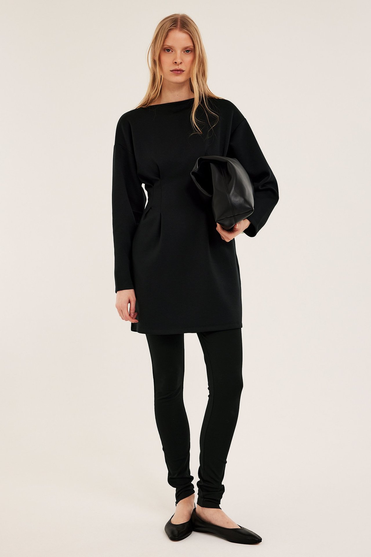 Framifrån: Kvinna i svart jerseyklänning med markerad midja, svarta leggings, platta skor, håller svart clutchväska.