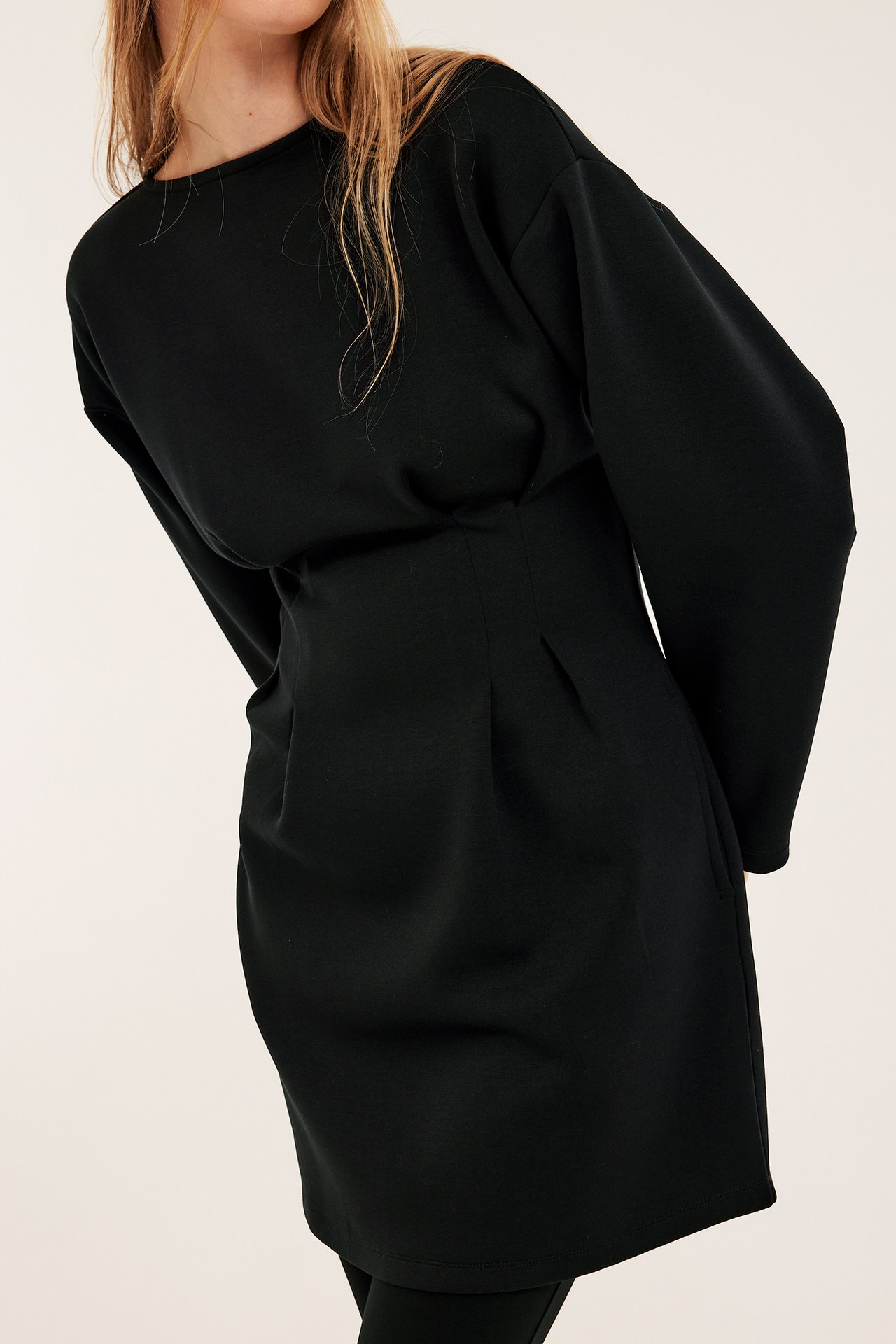 Från sidan, kvinna i svart enfärgad jerseyklänning med intagen midja och sidfickor.
