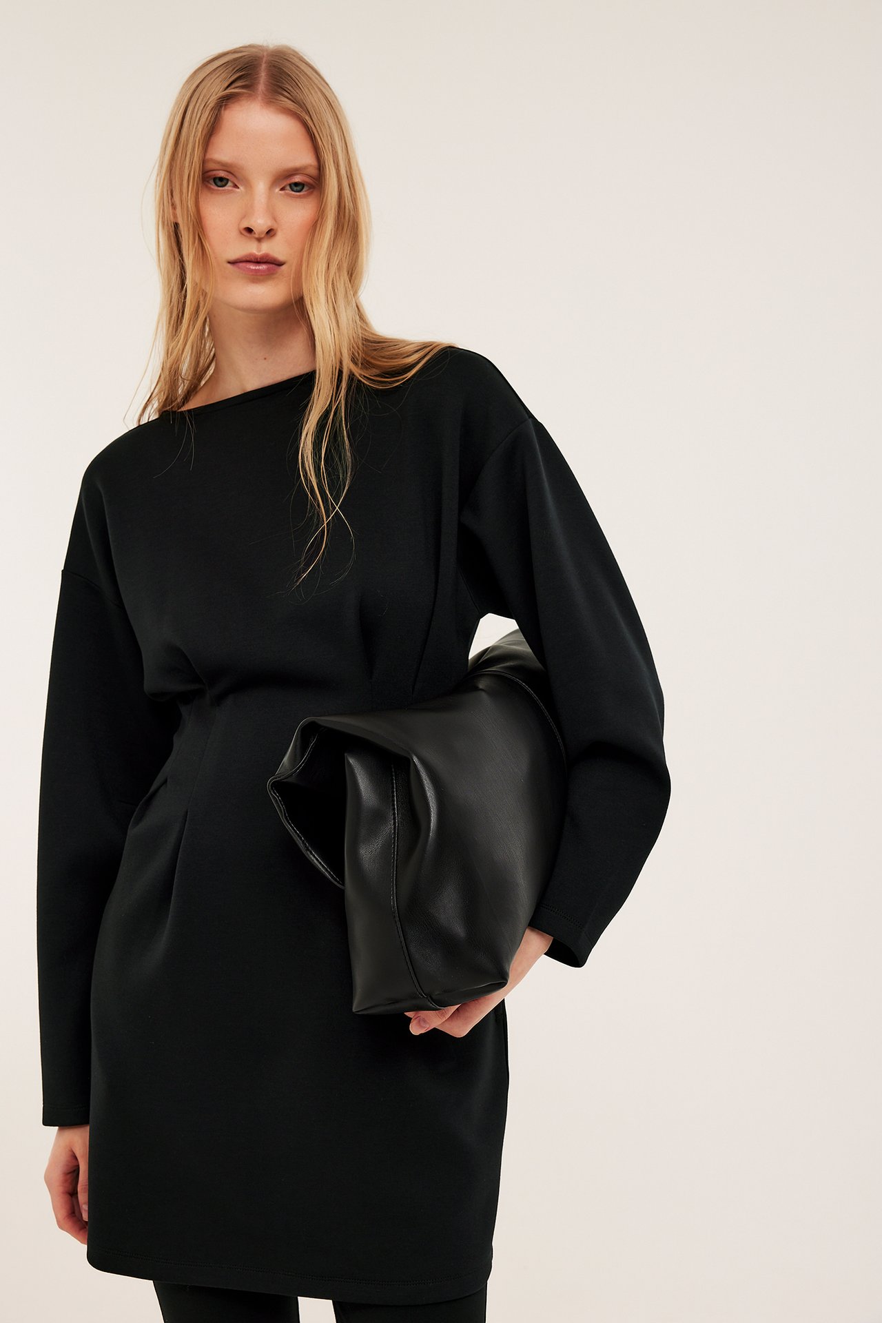 Frontbilde av kvinne i svart jerseykjole med markert midje, holdende en svart clutchveske.