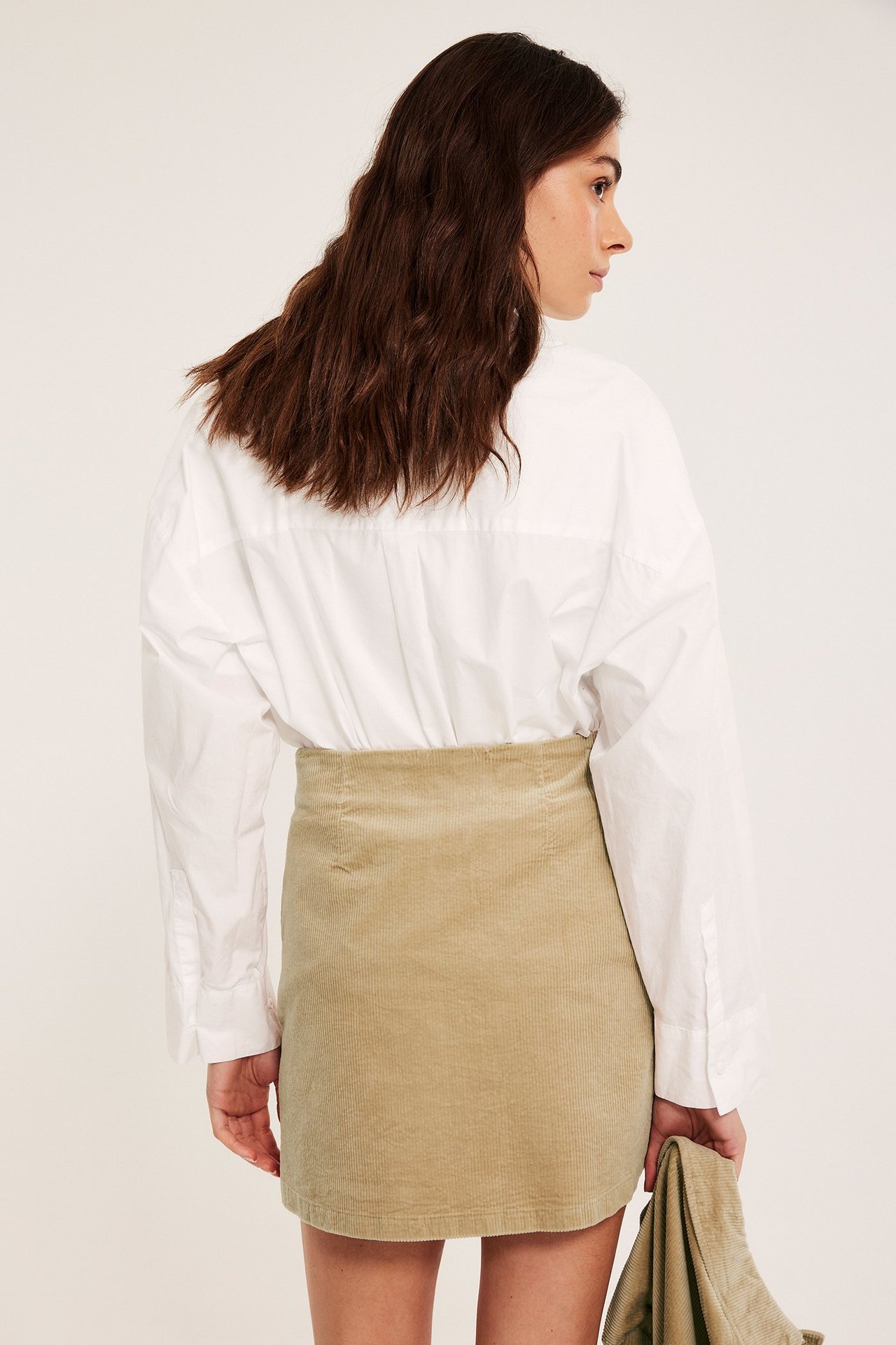 Widok z tyłu kobiety w beżowej sztruksowej mini spódnicy, białej koszuli, trzymającej pasującą kurtkę.