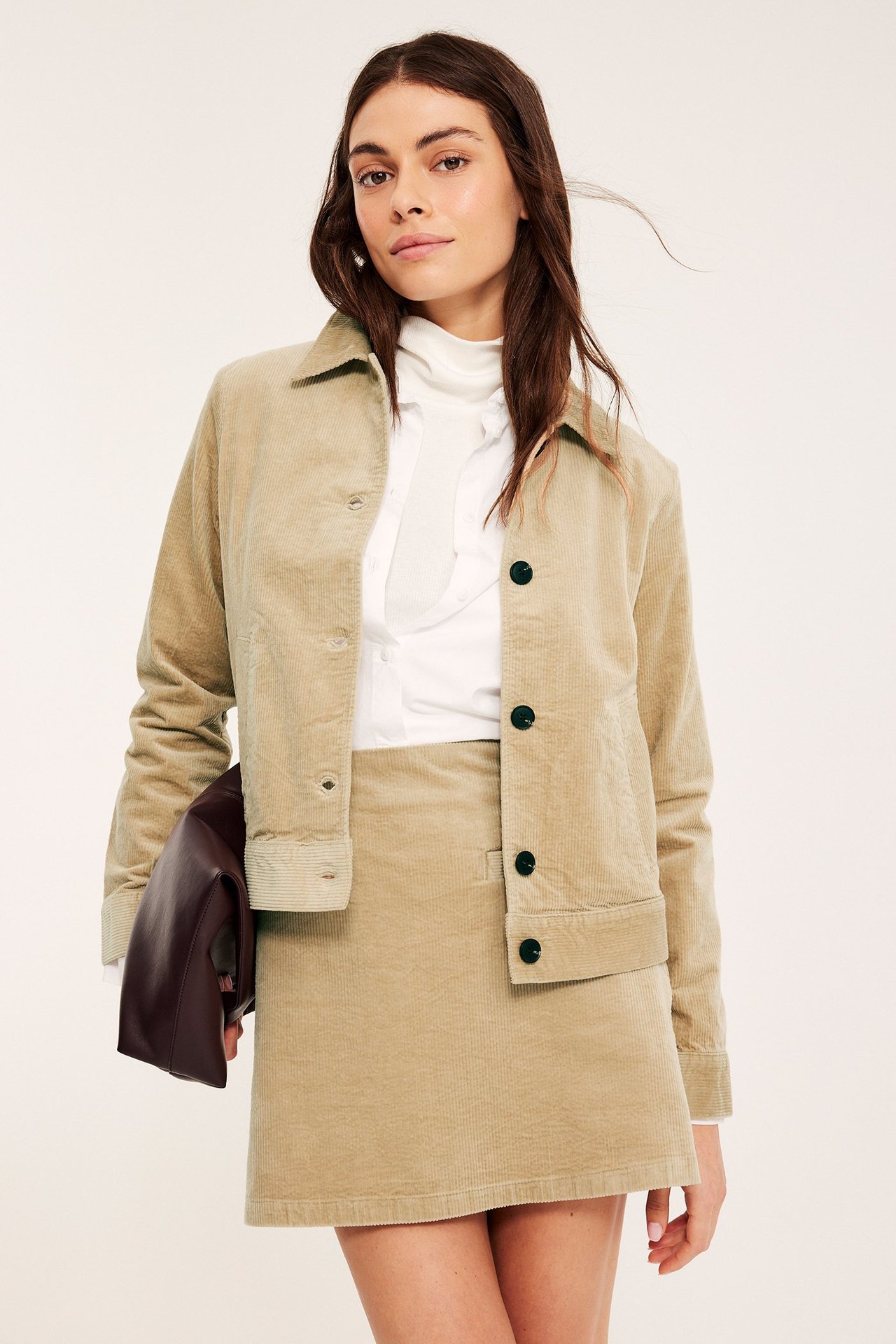 Edestä: Nainen beigessä samettihameessa, yhteensopivassa takissa, valkoisessa topissa, viininpunainen clutch.