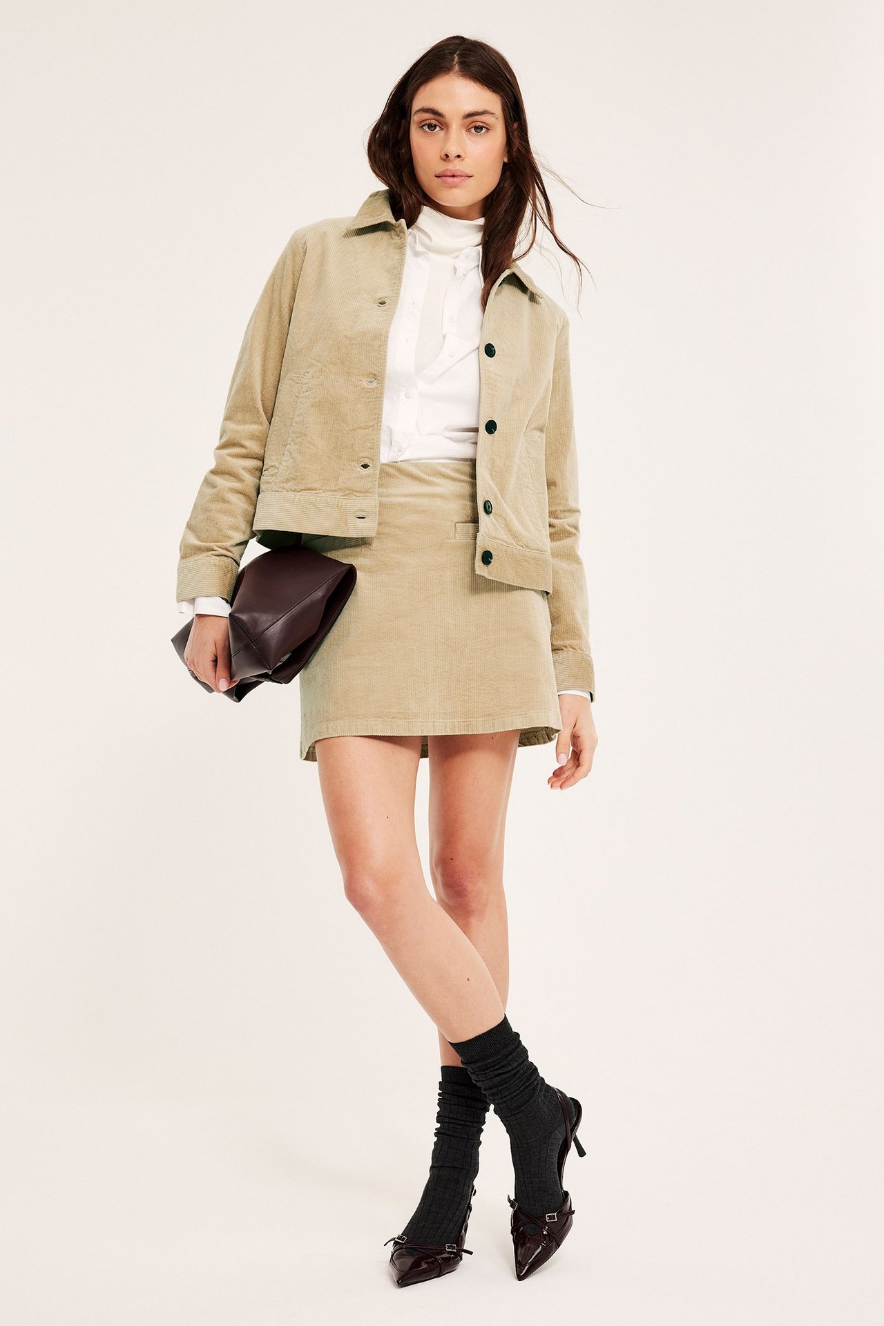 Widok z przodu kobiety w krótkiej beżowej spódnicy sztruksowej, kurtce, białej bluzce i brązowej kopertówce.
