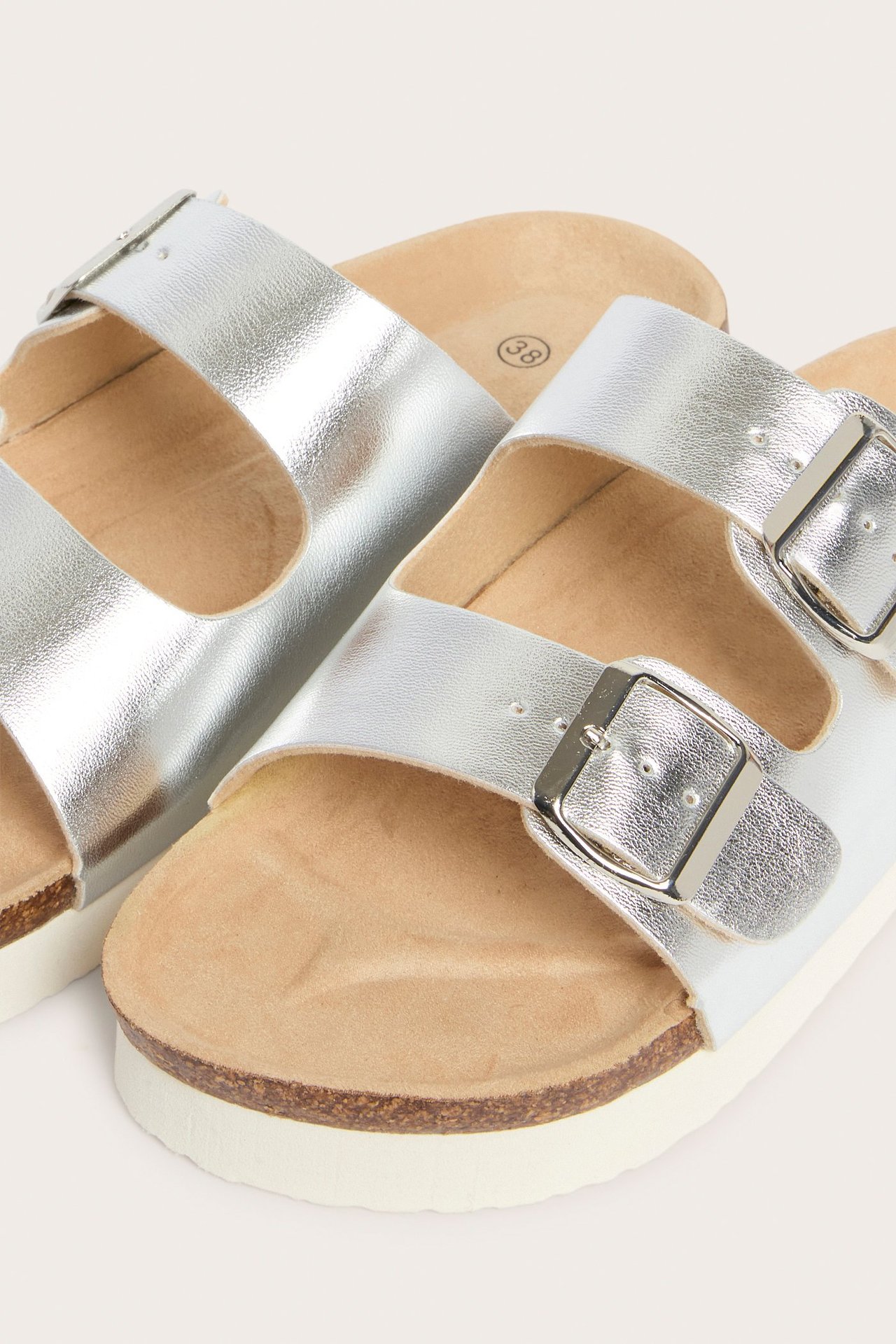 Lähikuva hopeisista PU-sandaaleista, joissa kaksi hihnaa, hopeasoljet ja korkkipohjat valkoisilla alustoilla.