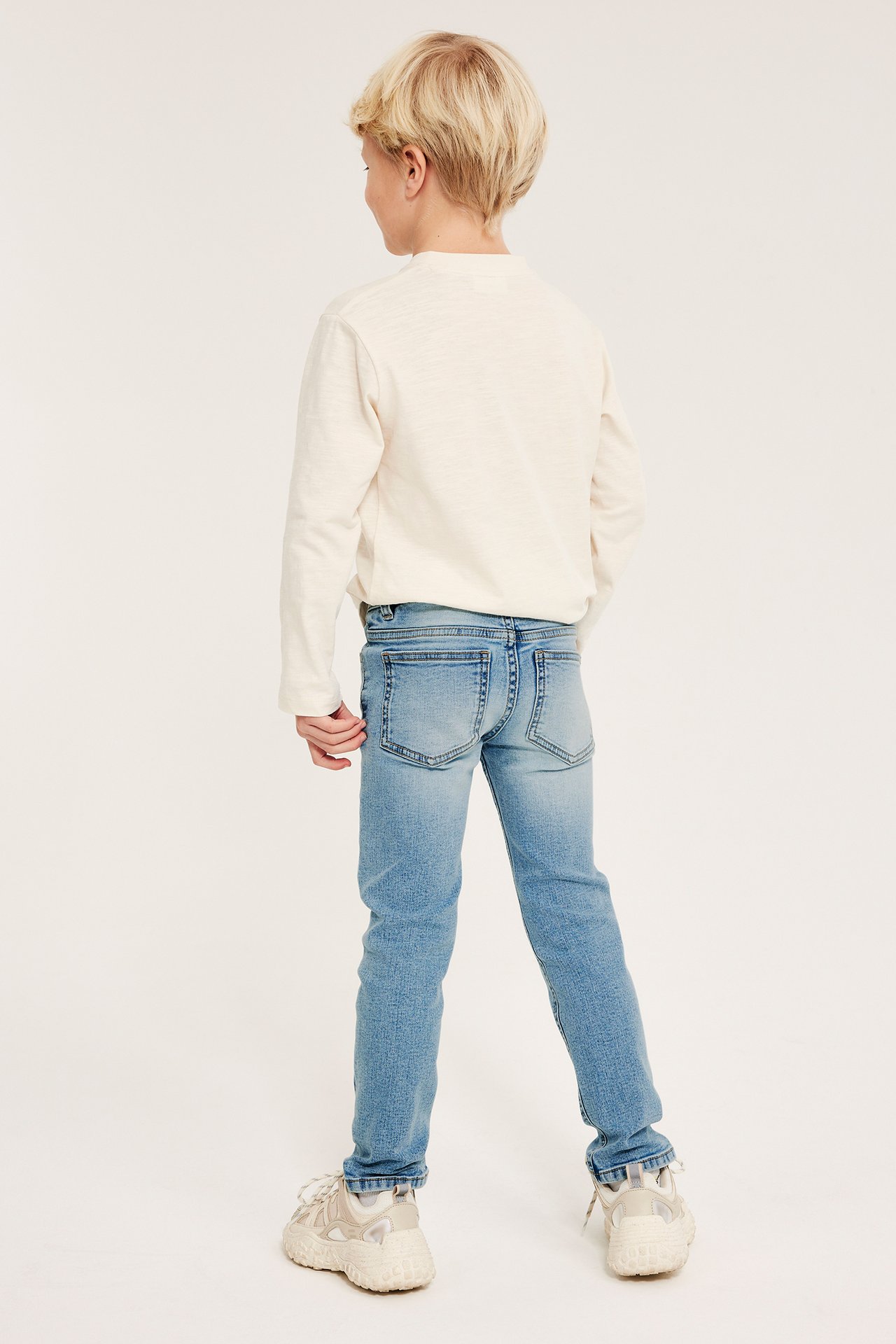 Bakifrån: Barn i ljusblå smala superstretch-jeans, gräddvit långärmad tröja och sneakers.