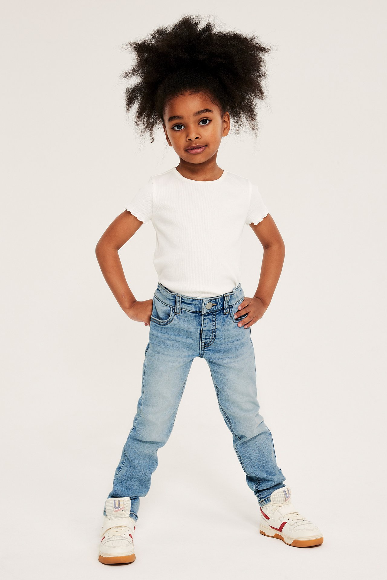 Framifrån, ett barn i ljusblå smala superstretch-jeans, en vit ribbad t-shirt och vita höga sneakers.