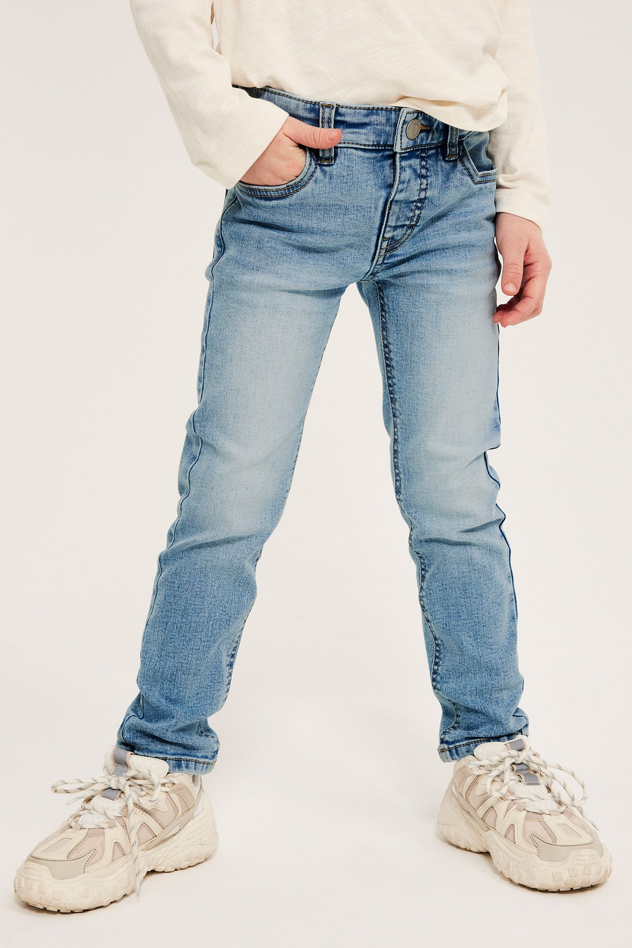 Framifrån, ett barn i ljusblåa slim-jeans, en krämvit långärmad tröja och beiga sneakers.