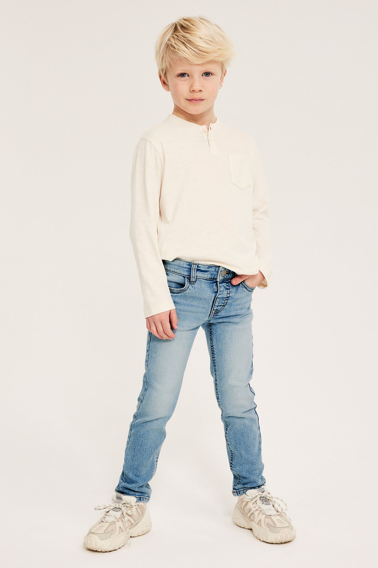 Framifrån: Pojke i ljusblå smala denimjeans, en benvit Henley-tröja och beige sneakers.