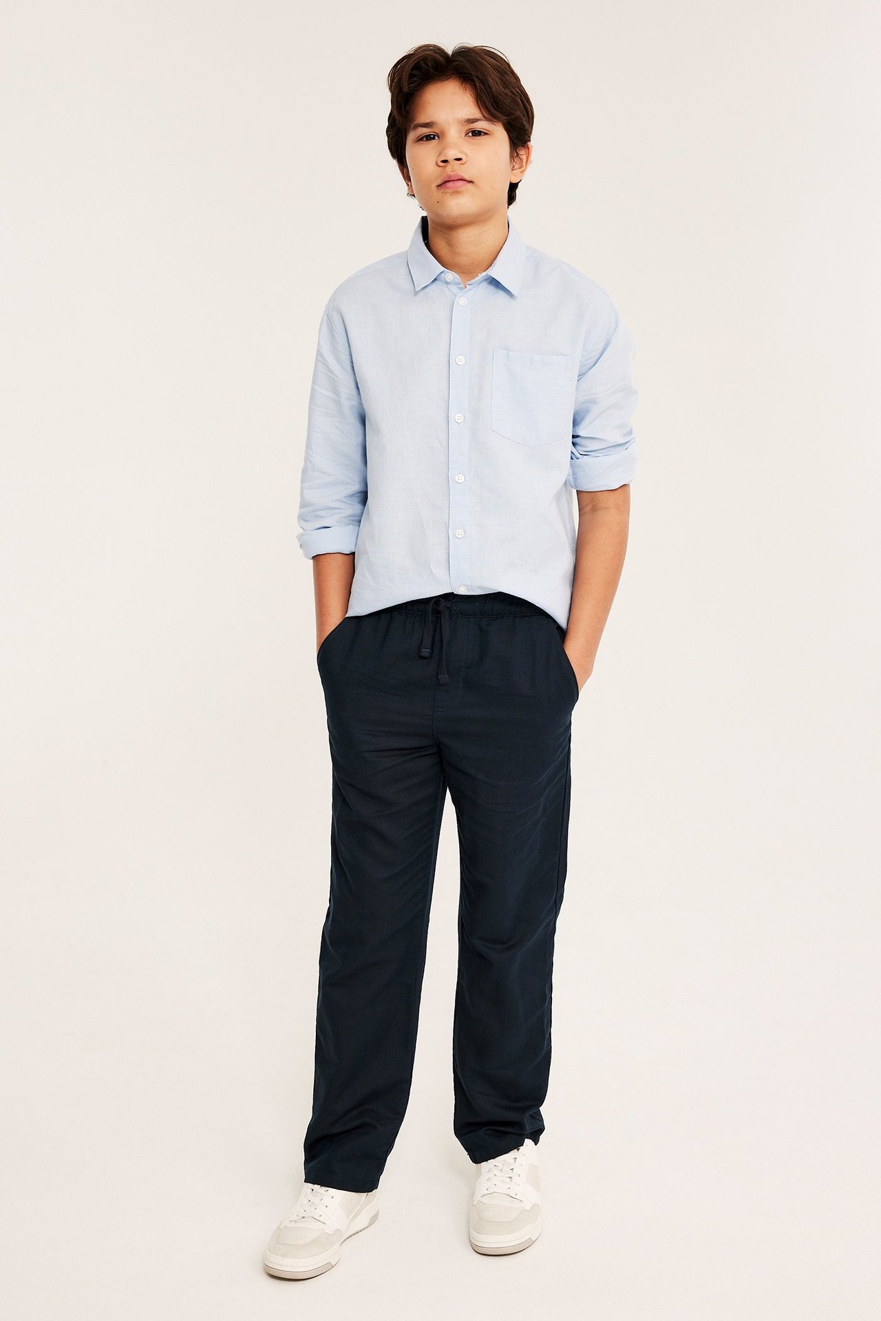 Edestä: Poika tummansinisissä pellavasekoitehousuissa, joissa kiristysnyöri, vaaleansininen paita ja valkoiset tennarit.