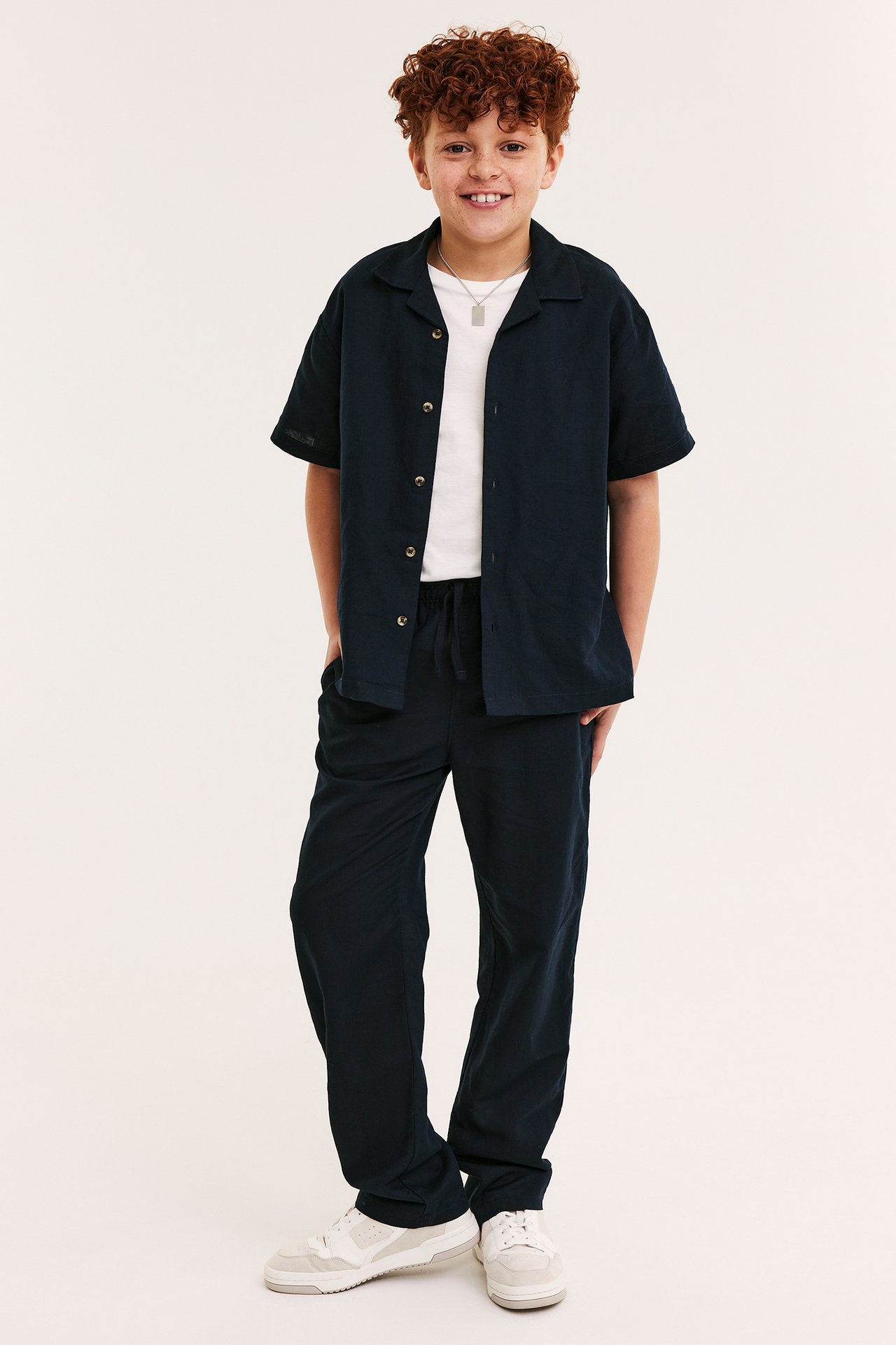 Framifrån: ett barn i mörkblå linnebyxor med dragsko, mörkblå skjorta över vit t-shirt, sneakers.