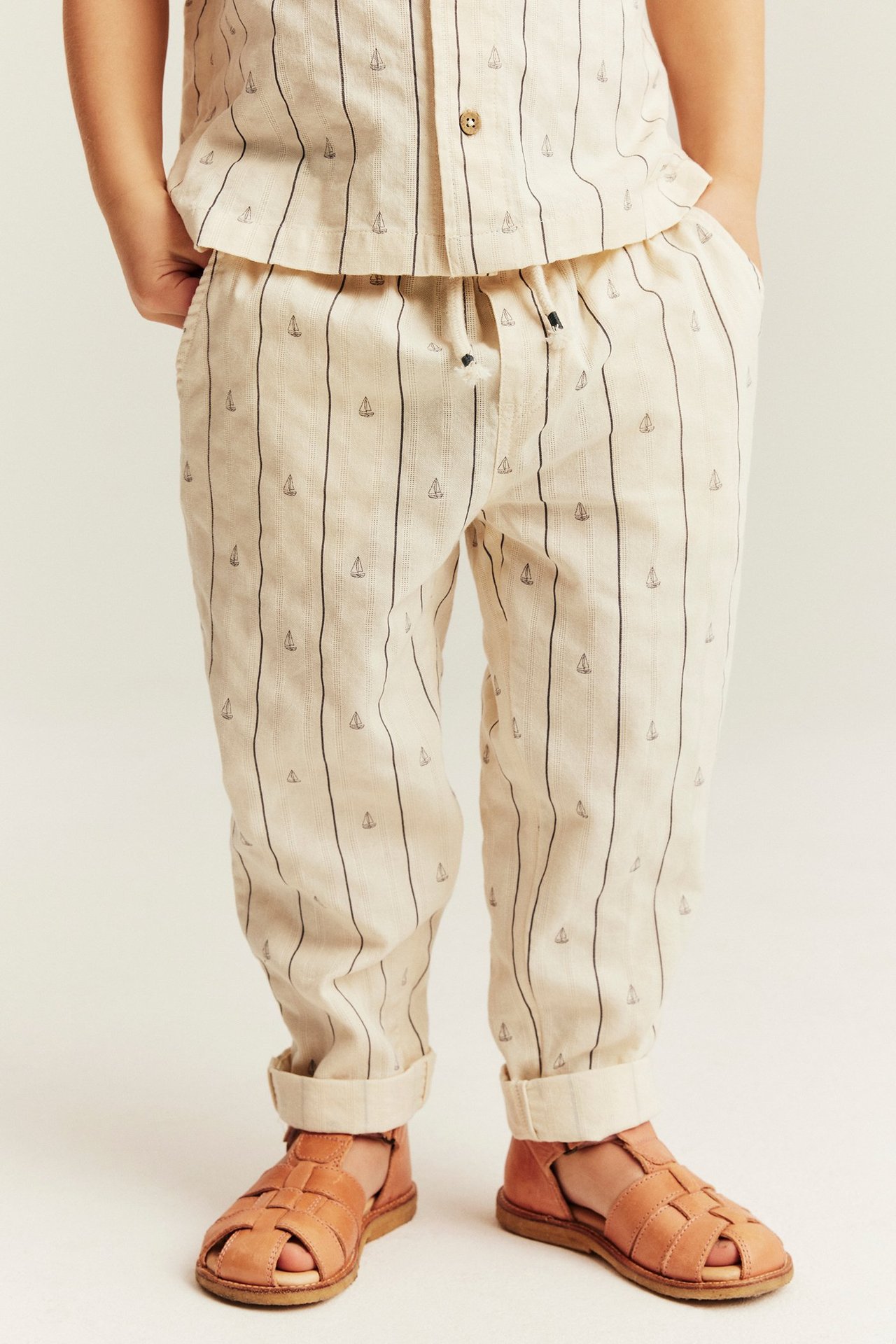 Frontvy av ett barn i offwhite randiga byxor med segelbåtar, matchande skjorta och bruna sandaler.