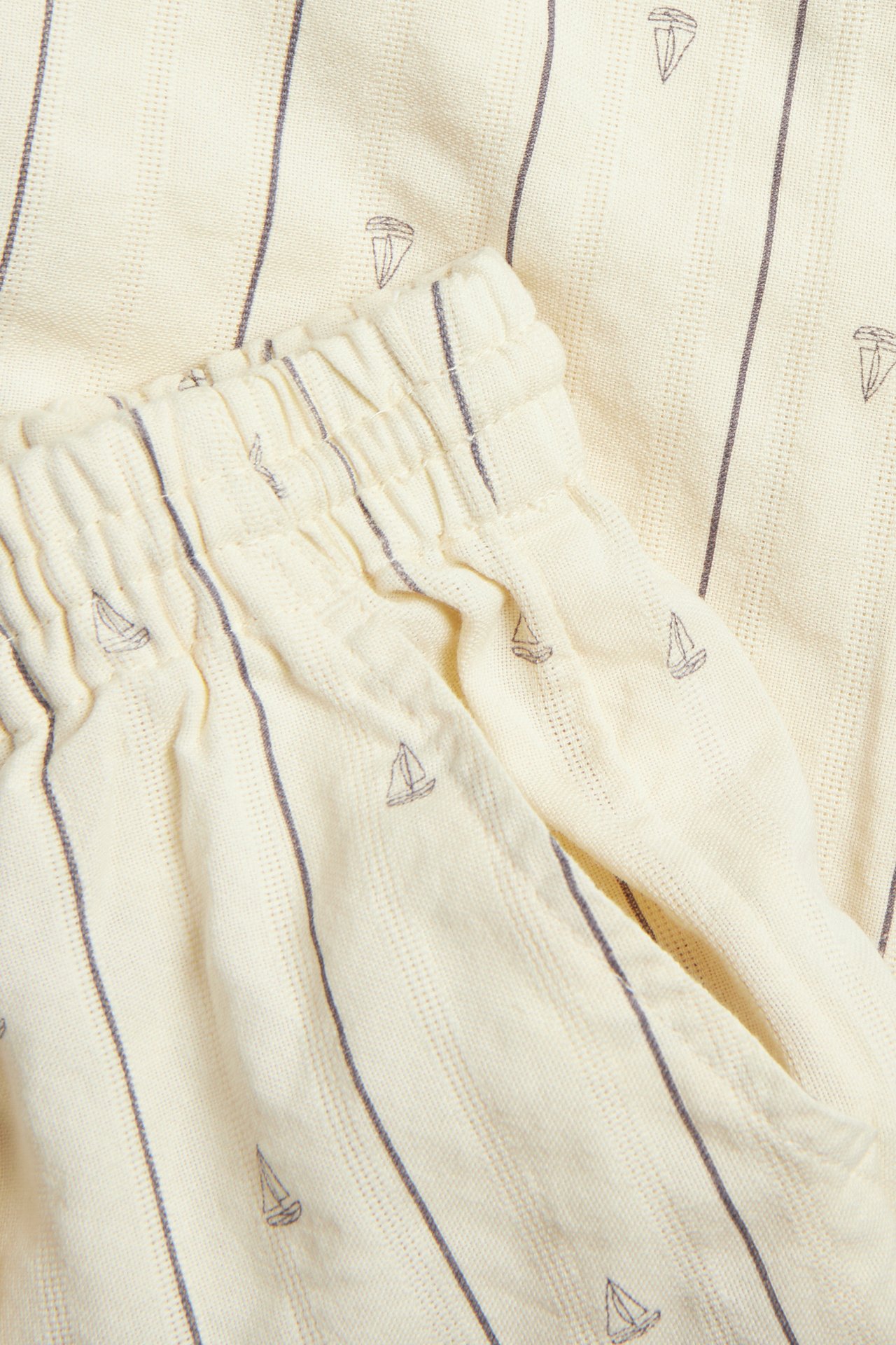 Lähikuva kermanvärisistä puuvillahousuista harmailla raidoilla ja purjevenekuvioilla, joustavalla vyötäröllä.