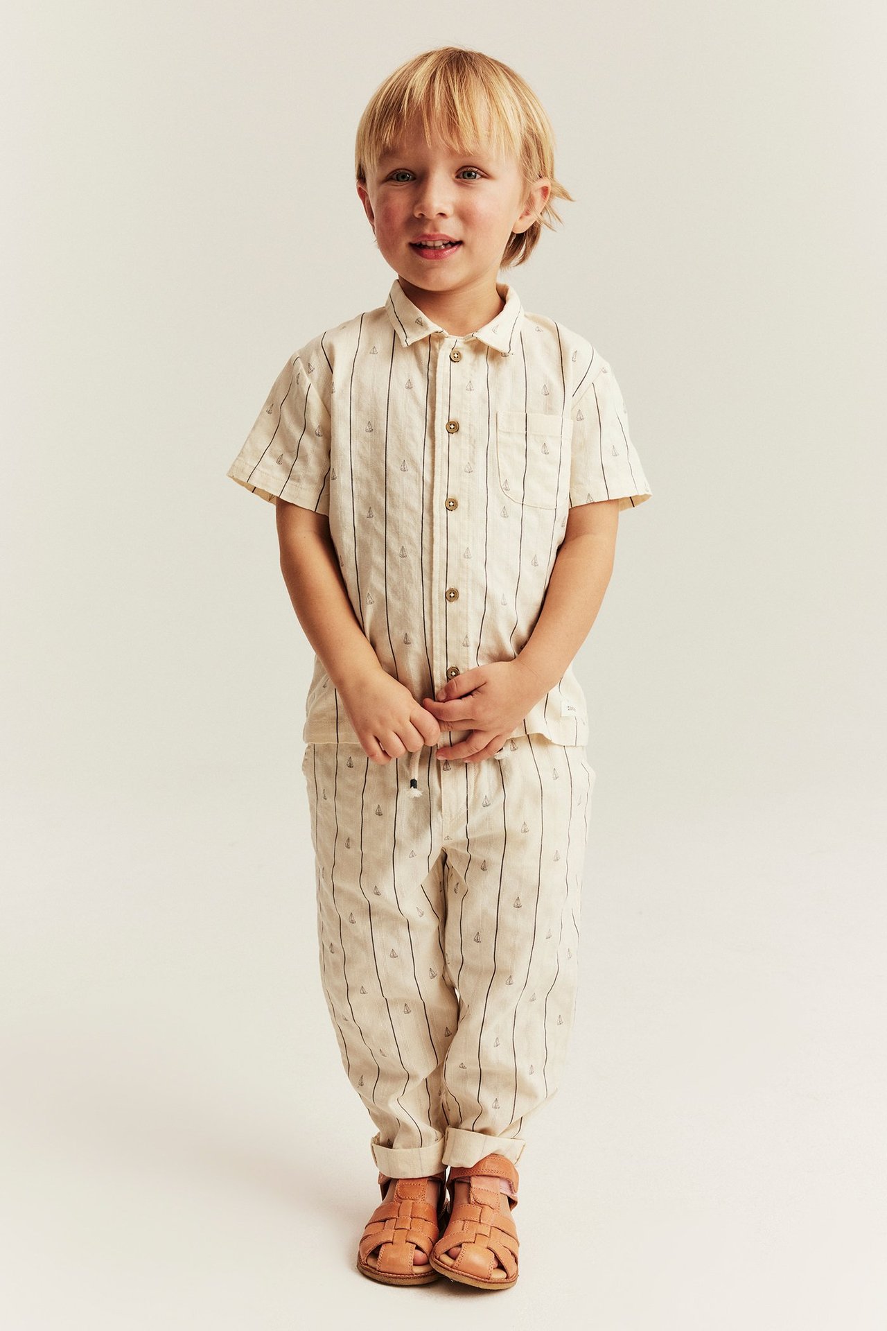 Widok z przodu: Dziecko w kremowych, bawełnianych spodniach w paski z żaglówkami, pasującej koszuli, sandałach.