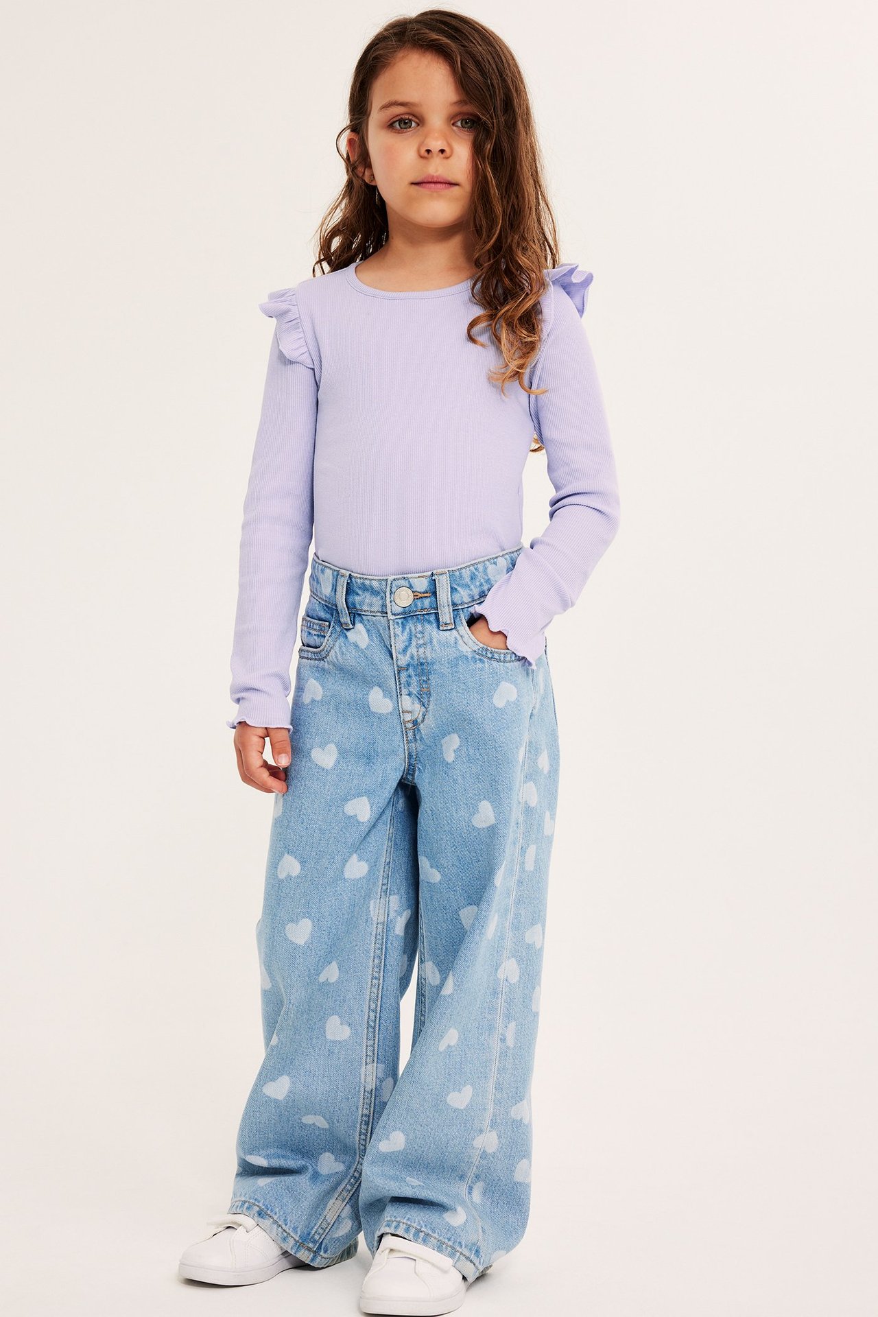 Framsida av ett barn i en ljuslila ribbad långärmad topp med volanger och hjärtmönstrade jeans.