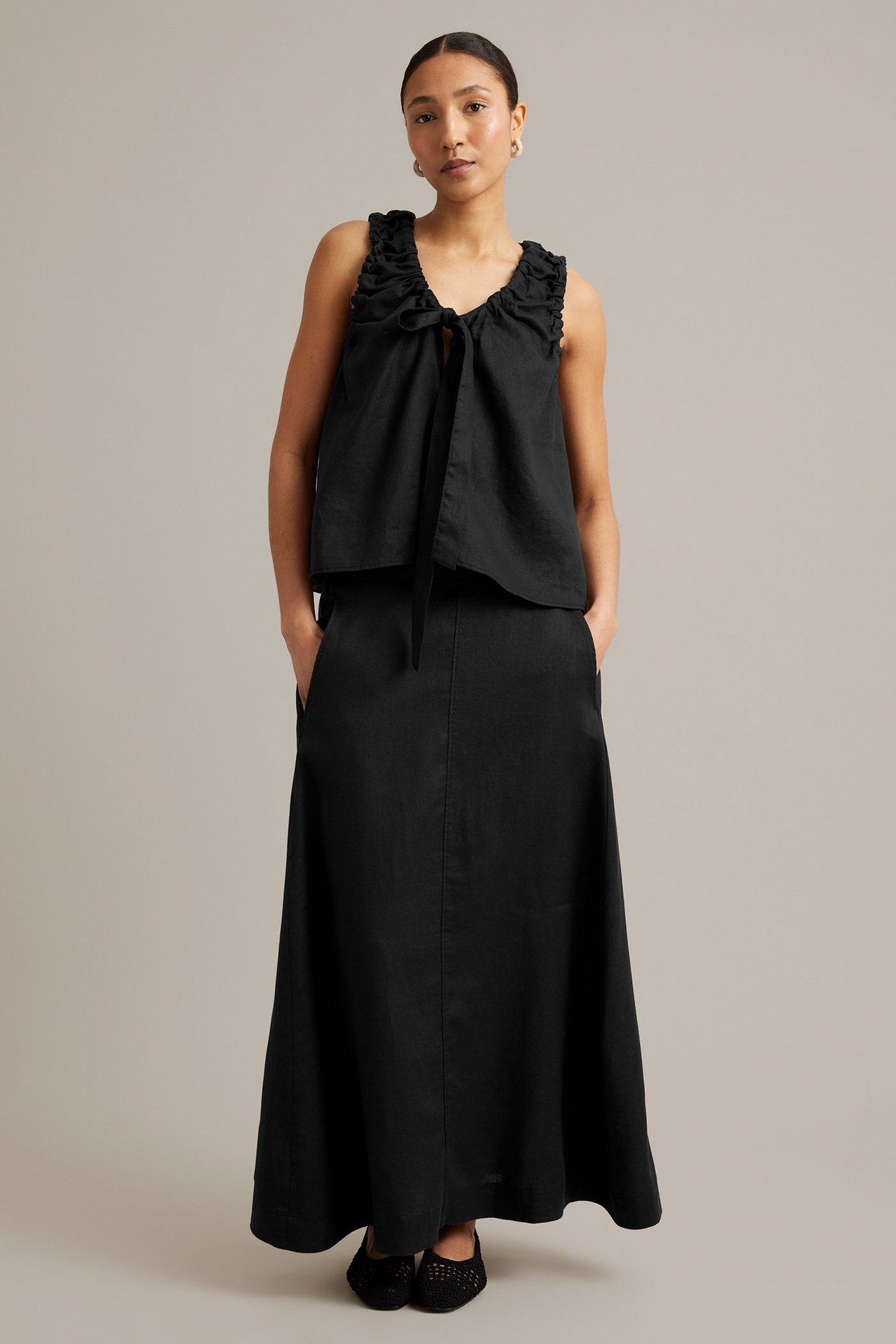 Hel framifrån: kvinna i svart linnetopp med rynkad knytrigning och en svart maxikjol.