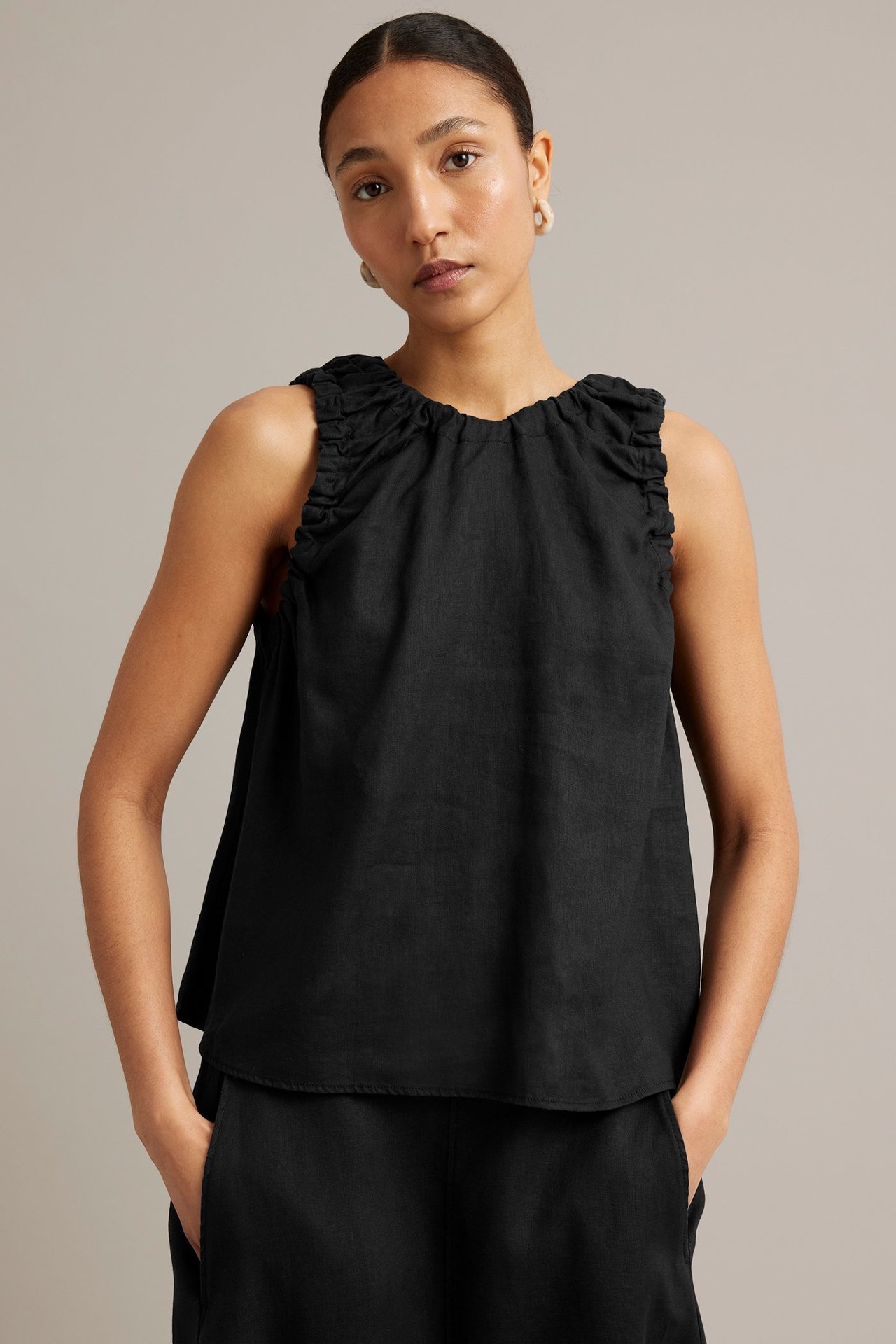 Framsida av kvinna i svart ärmlös topp i linne med rynkad halsringning, svarta byxor och ljusa örhängen.